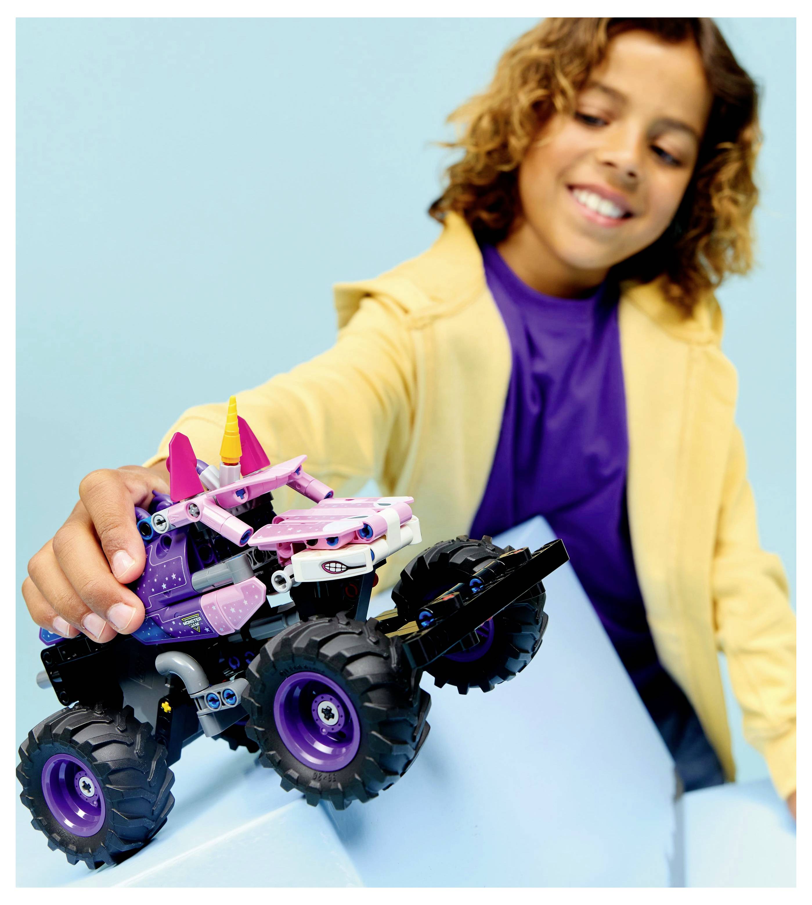 Ein Kind in einer gelben Jacke und einem lila Shirt lächelt, während es mit einem bunten Spielzeug-Monstertruck mit großen Rädern und lebhaften Farben spielt.