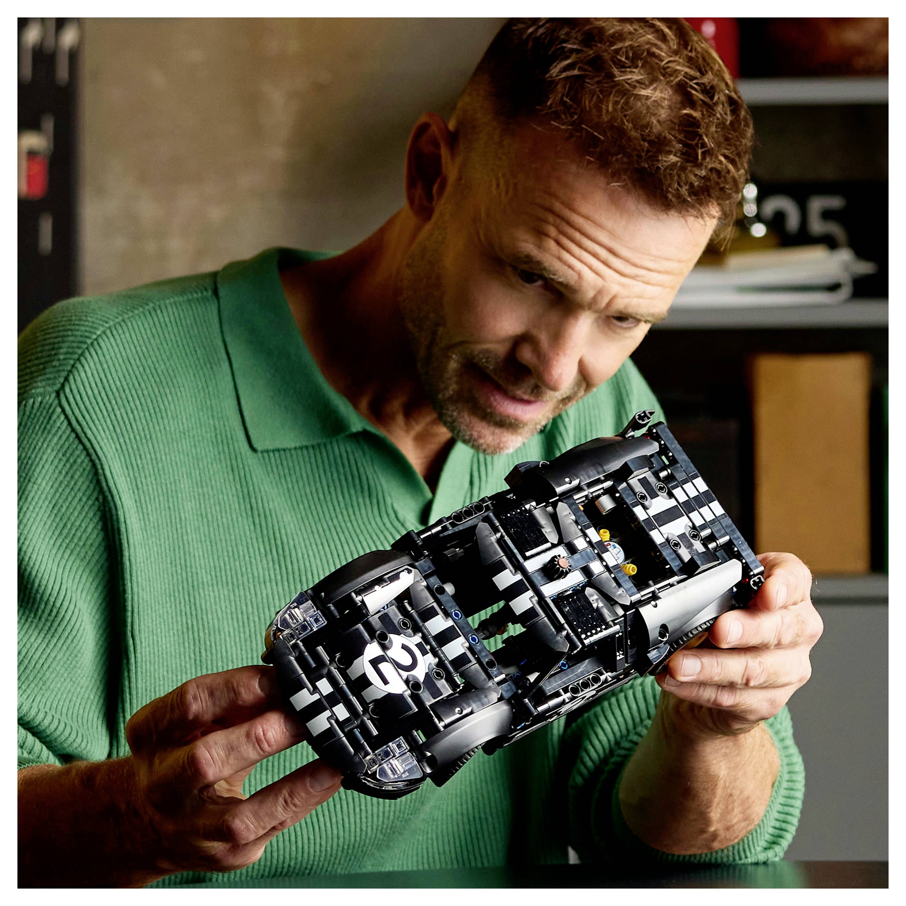 Mann in grünem Pullover, der ein schwarzes Modelrennfahrzeug mit der Nummer '25' in einer garagenähnlichen Umgebung untersucht.