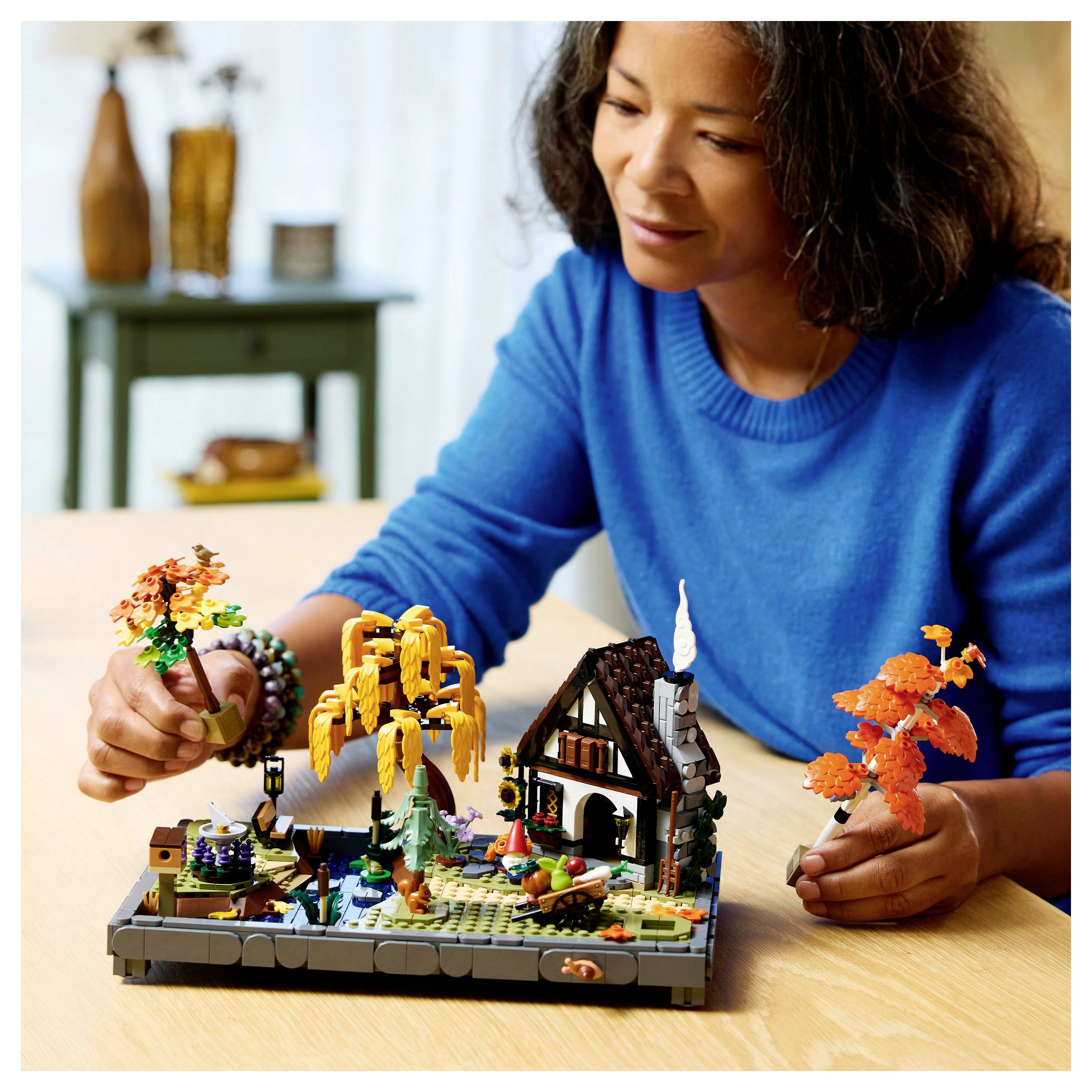 Eine Person in einem blauen Pullover baut eine detaillierte Herbst-Szene aus LEGO auf einem Tisch, mit Bäumen und einem kleinen Haus.