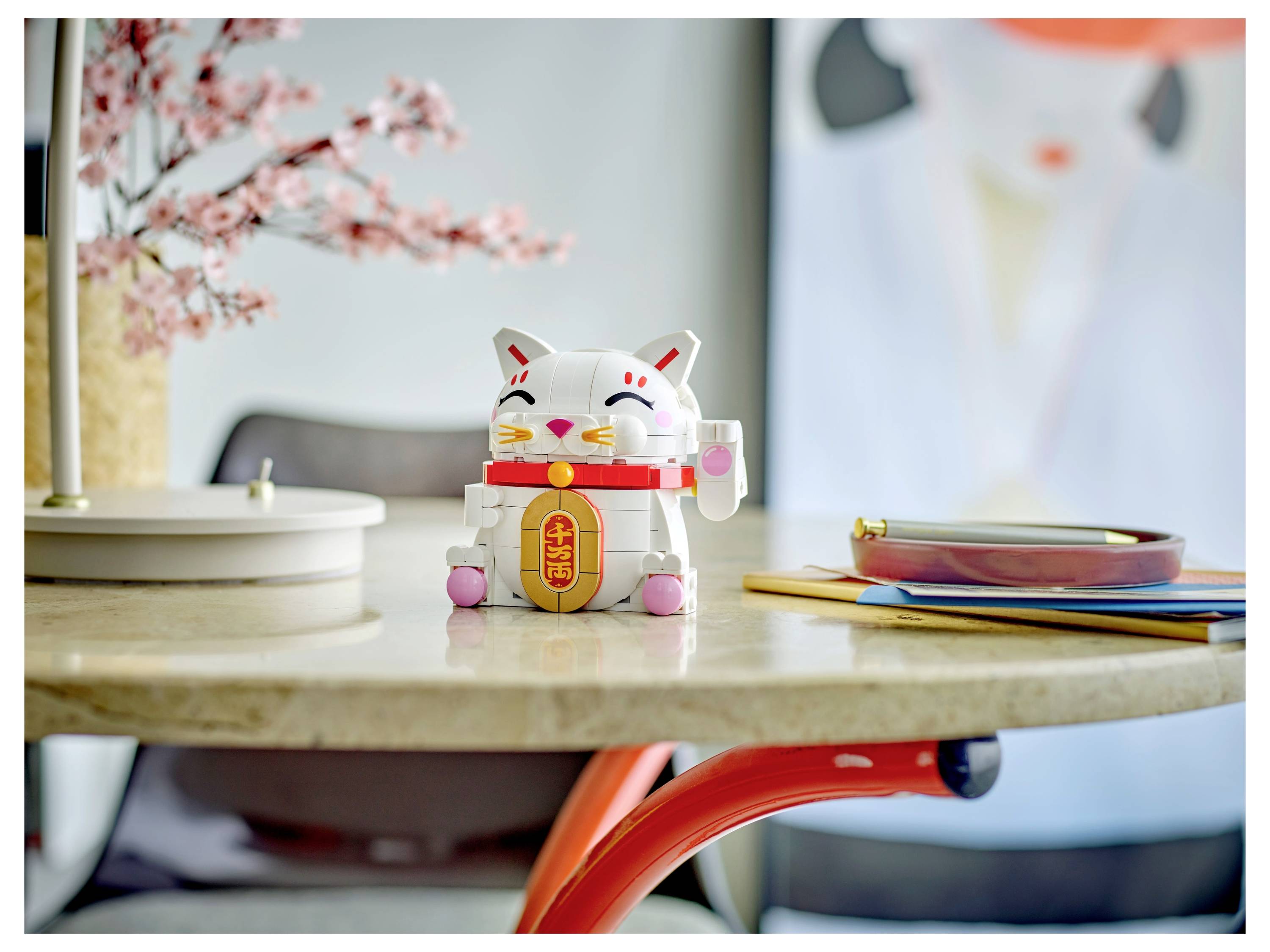 Eine Katzen-Keramikfigur mit gehobener Pfote sitzt auf einem runden Tisch neben Kirschblüten und Notizbüchern und symbolisiert gutes Glück.