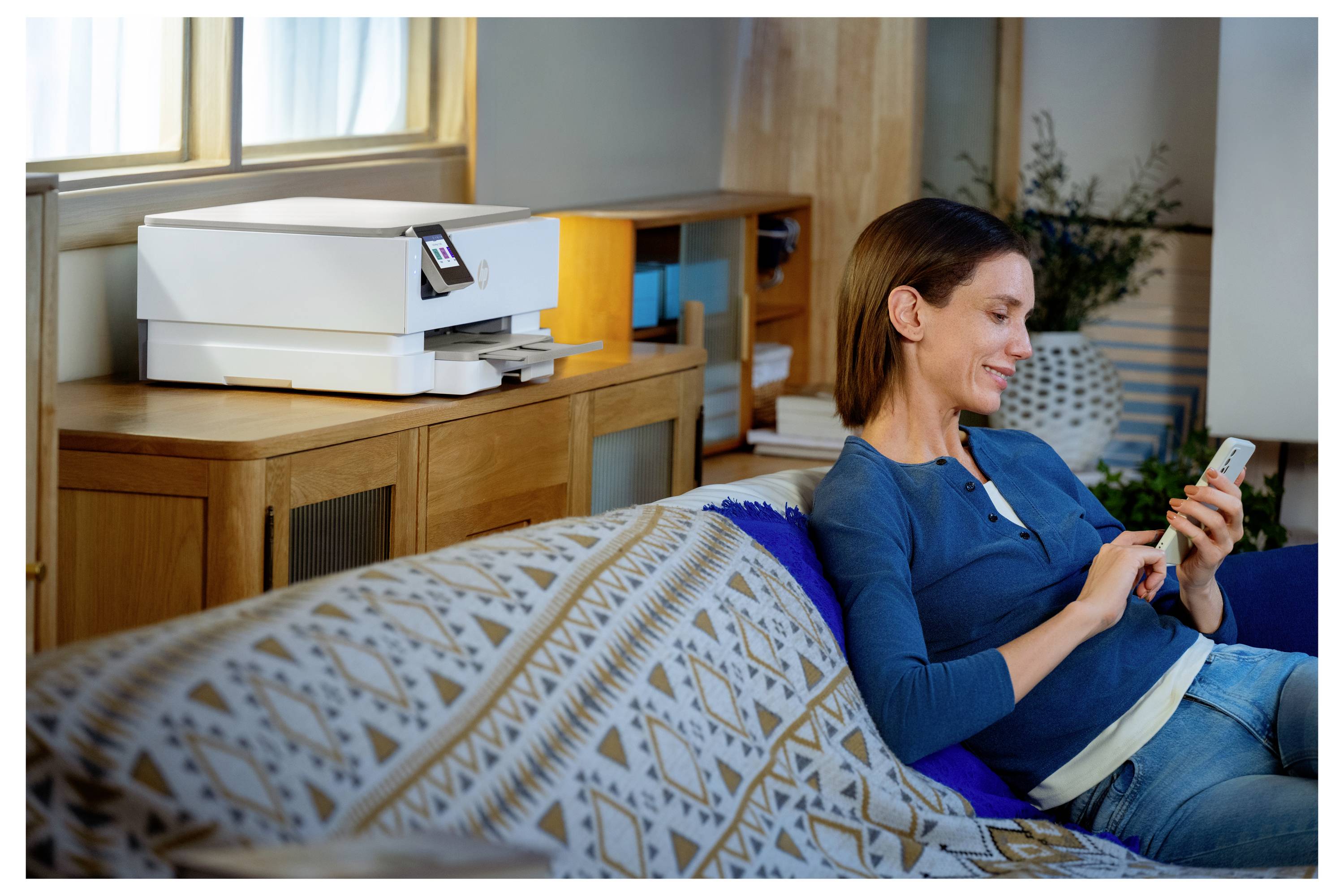 Eine Frau sitzt auf einem Sofa und lächelt auf ihr Smartphone. Ein moderner Drucker steht in einem Regal im Hintergrund, was eine entspannte Heimbüro-Atmosphäre andeutet.