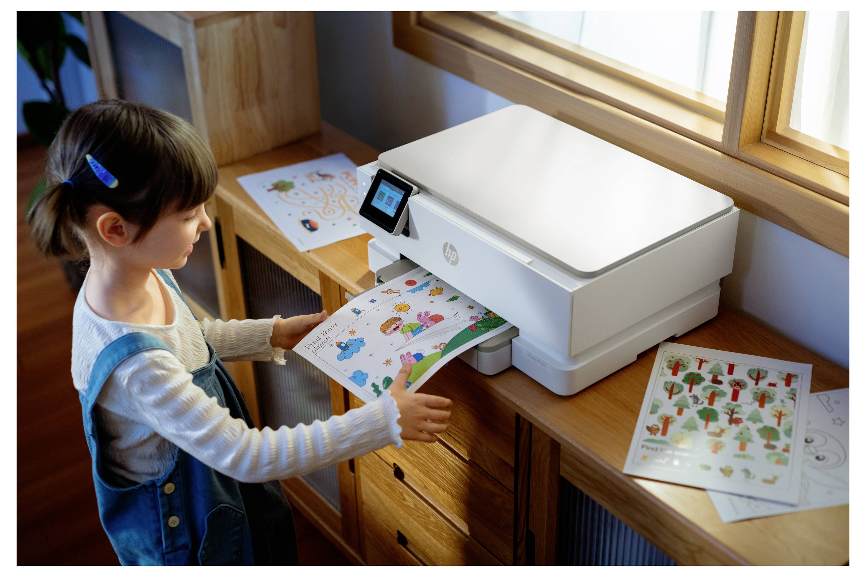 Ein Kind steht neben einem Drucker und holt farbenfrohe bedruckte Blätter mit verschiedenen Illustrationen heraus. Kunststoffmaterialien sind auf dem Tisch in der Nähe verstreut.