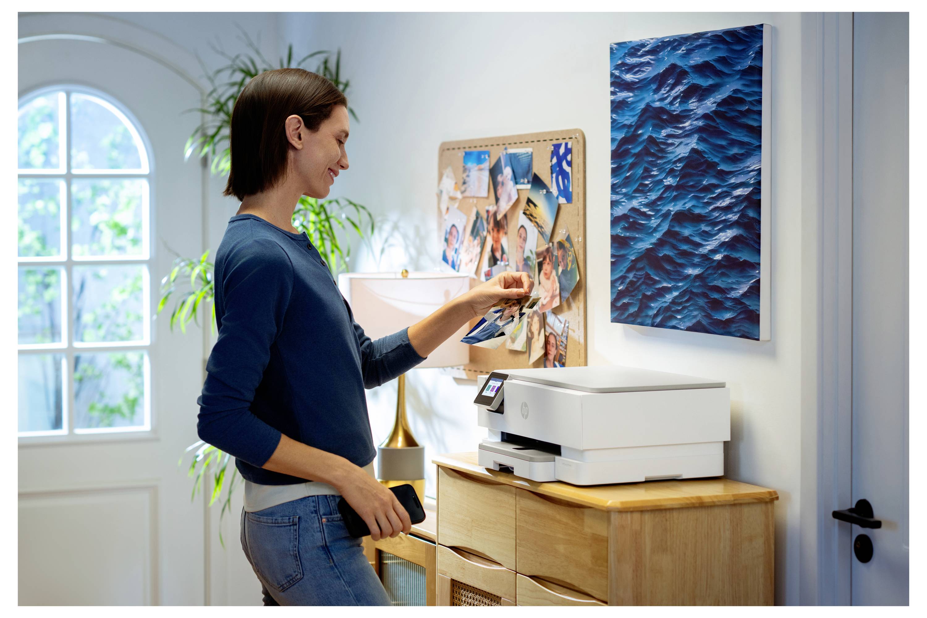 Eine Person legt ein Foto von einem Smartphone auf einen Heimdrucker, während sie in einem hell erleuchteten Raum steht, in dem eine Pinnwand und ein Gemälde in der Nähe hängen.