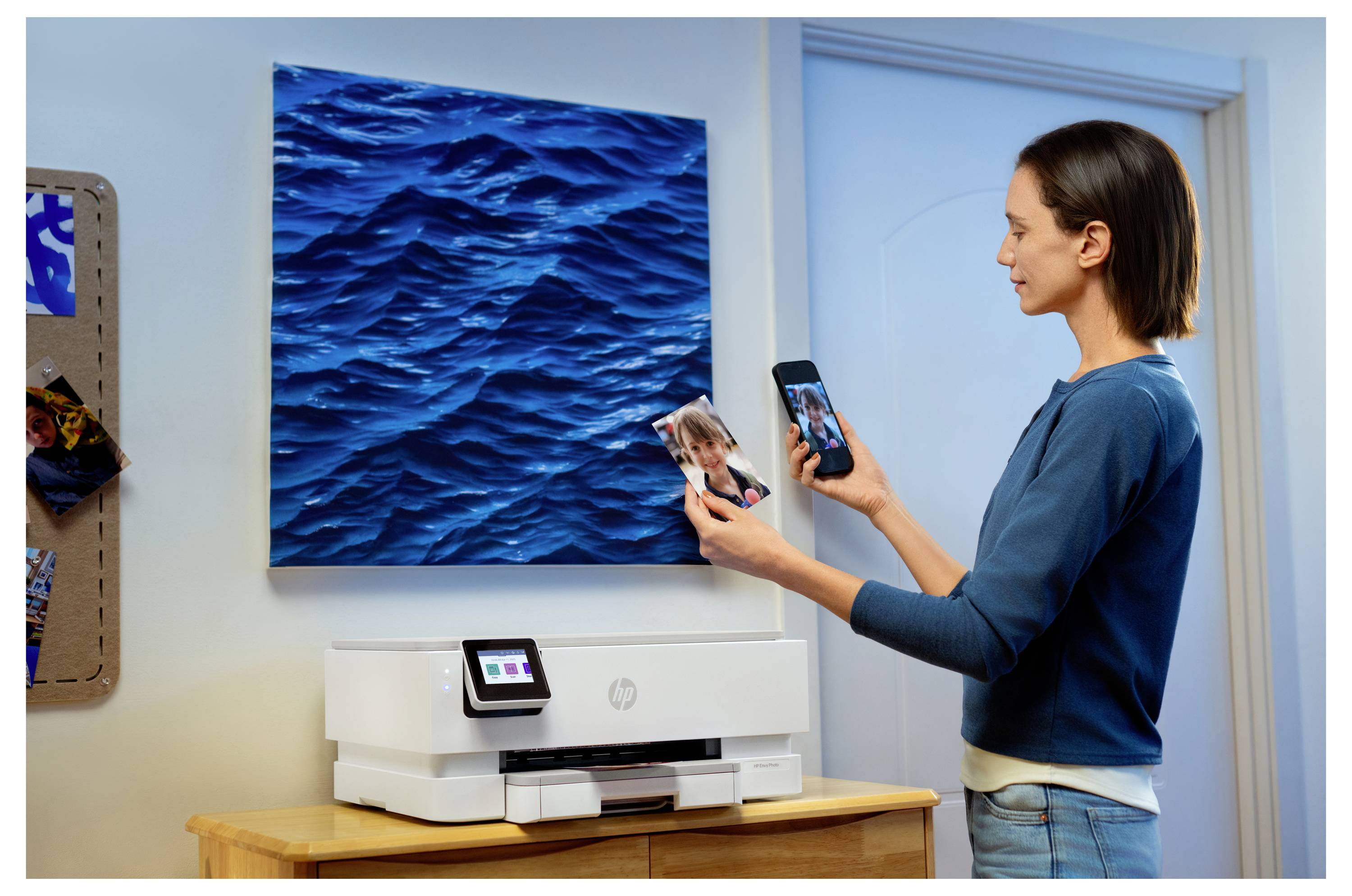 Frau hält zwei Fotos an ein Smartphone und vergleicht Bilder neben einem Drucker und einem blauen abstrakten Wanddruck.