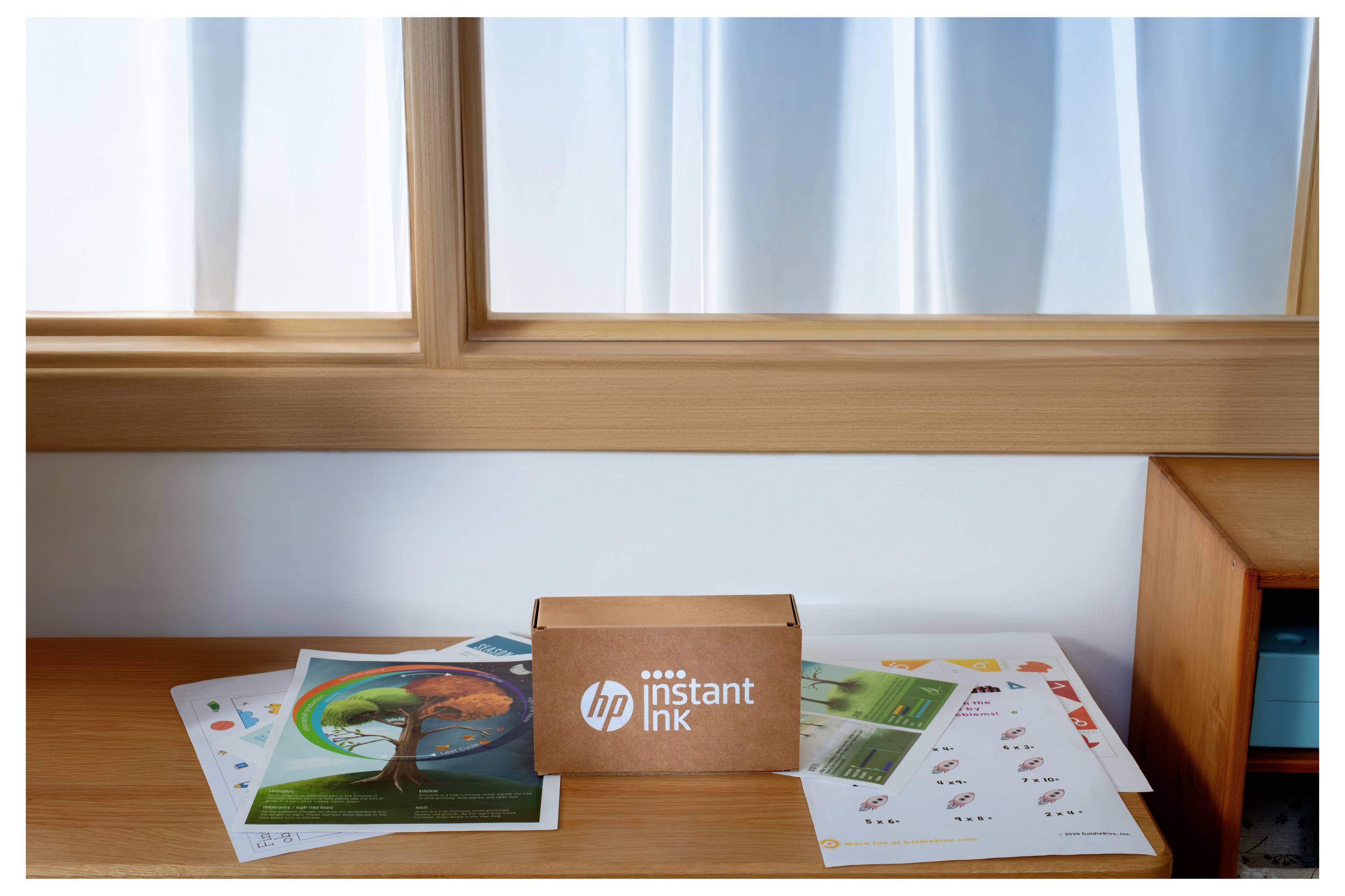 HP Instant Ink-Box auf einem Holztisch, umgeben von bunten Druckpapieren, neben einem Fenster mit weißen Gardinen.