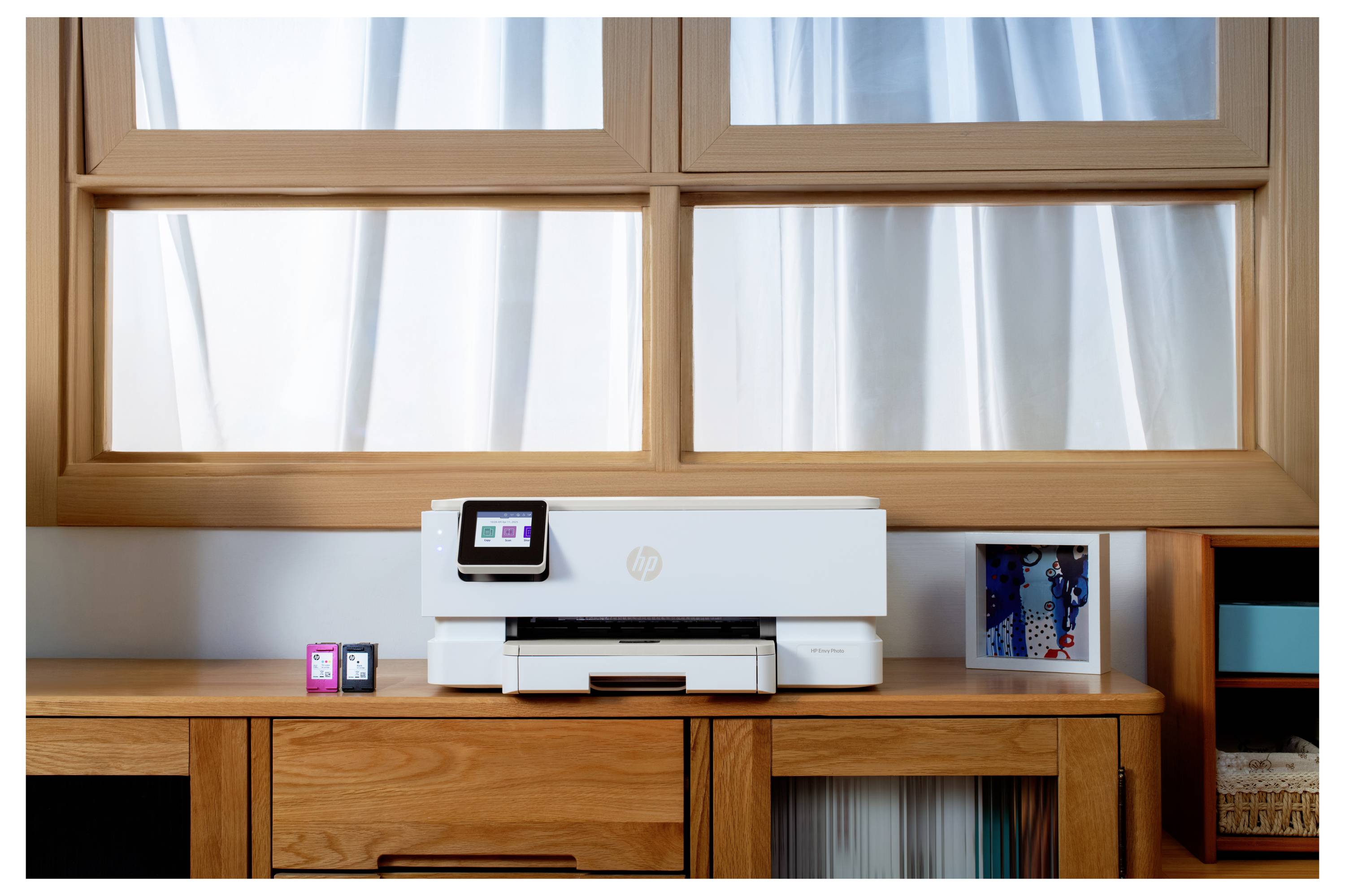 Ein Drucker steht auf einem Holzschreibtisch unter einem großen Fenster, mit zwei Tintenpatronen daneben und einem kleinen gerahmten Bild rechts davon.