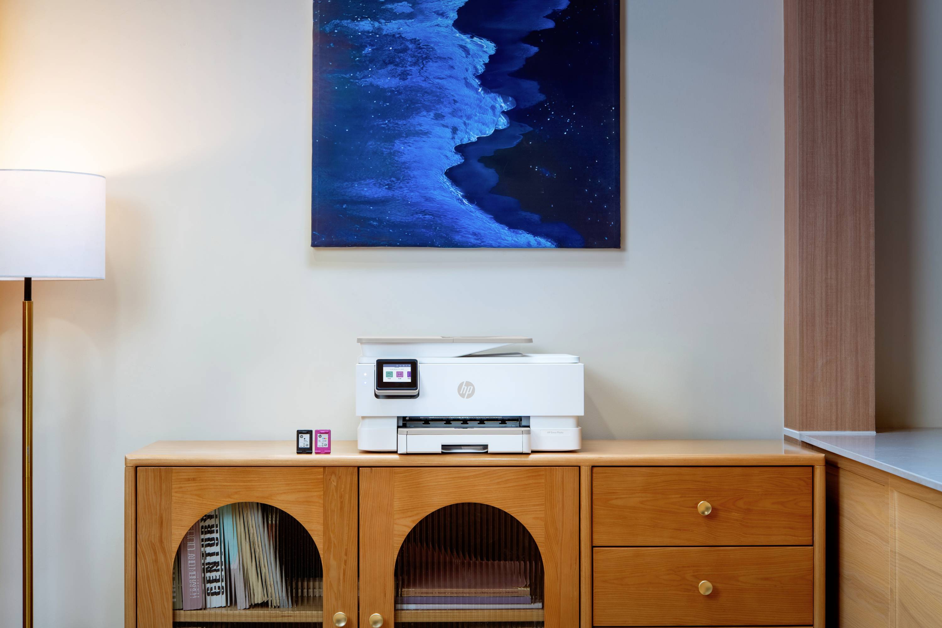 Moderner Drucker auf einem hölzernen Sideboard in einem minimalistischen Zimmer, mit einer Stehlampe und blauem Ozeankunstwerk darüber.