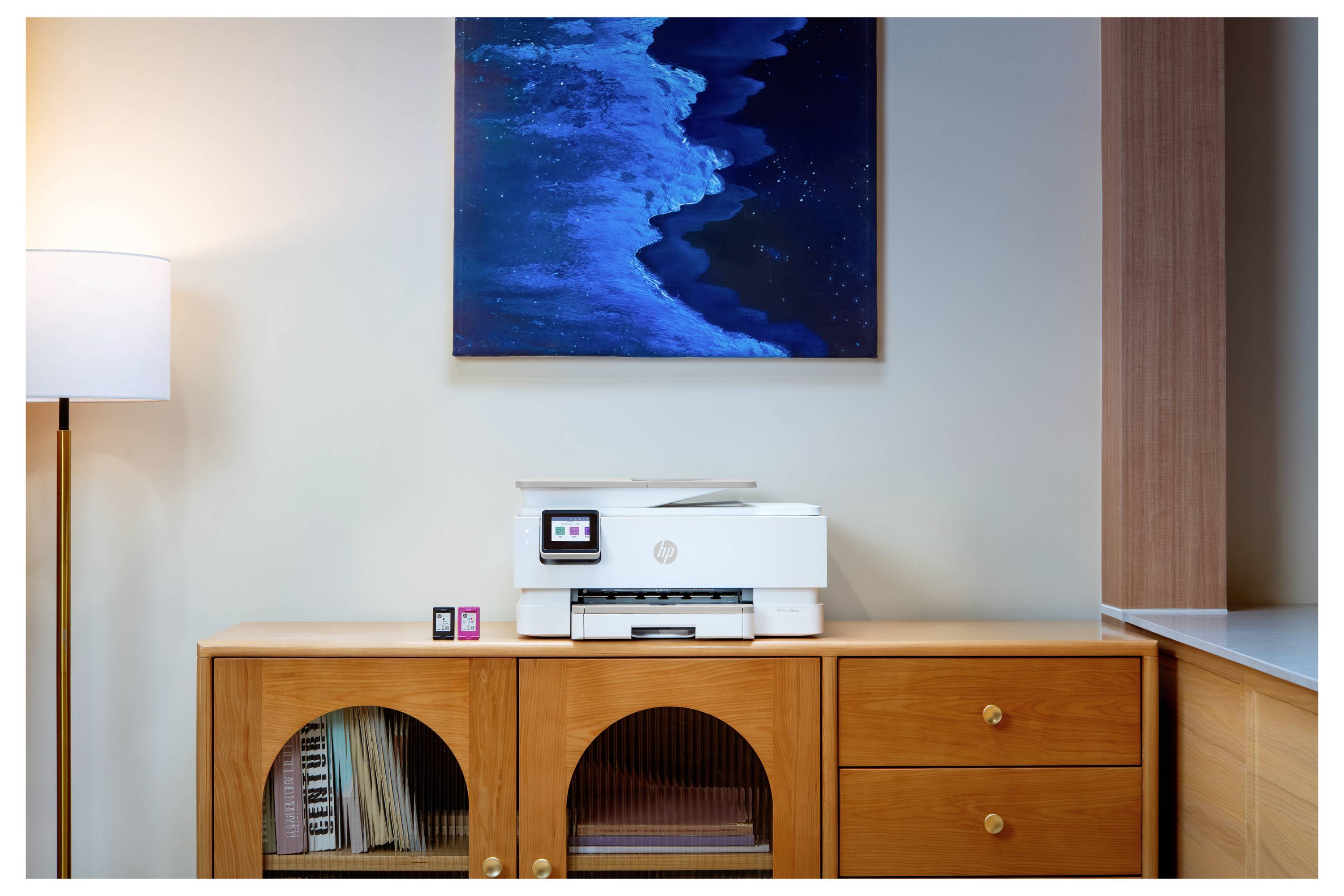 Ein hölzernes Sideboard mit einem Drucker und kleinen Tintenpatronen unter einem blauen abstrakten Wandgemälde, neben einer Stehleuchte.