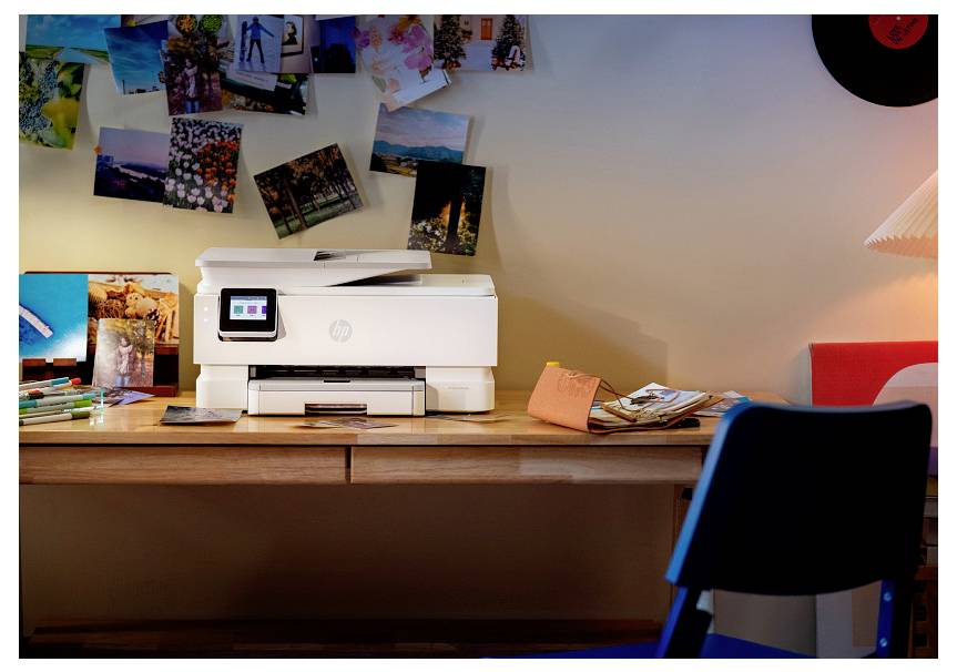 Schreibtisch mit einem HP-Drucker in der Mitte auf einem Holztisch, umgeben von verstreuten Schreibwaren, Postkarten und Fotos, die an der Wand dahinter angeheftet sind.