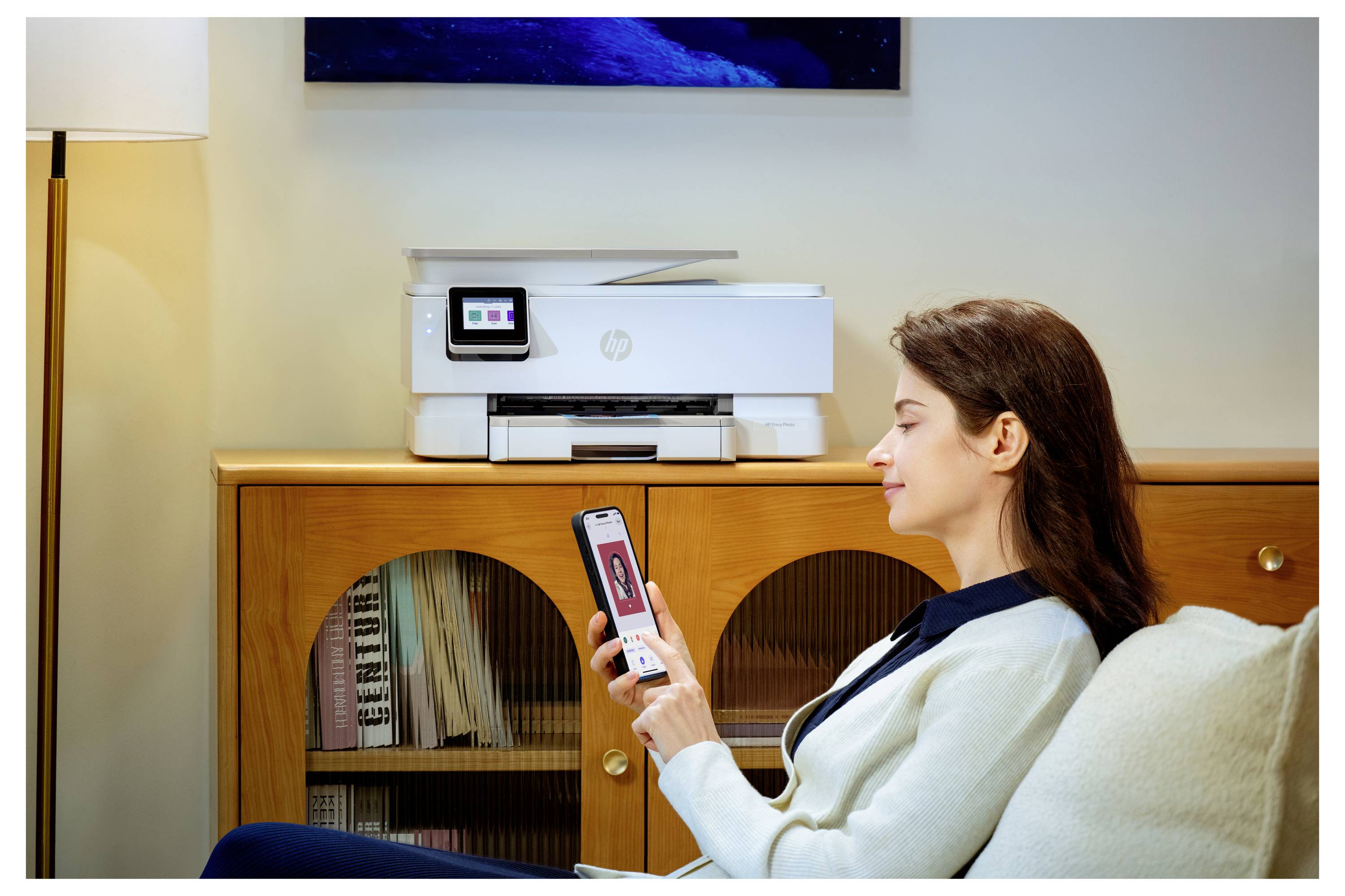 Eine Frau sitzt auf einem Sofa und benutzt ein Smartphone neben einem HP-Drucker auf einem Holzschrank in einem Wohnzimmer.