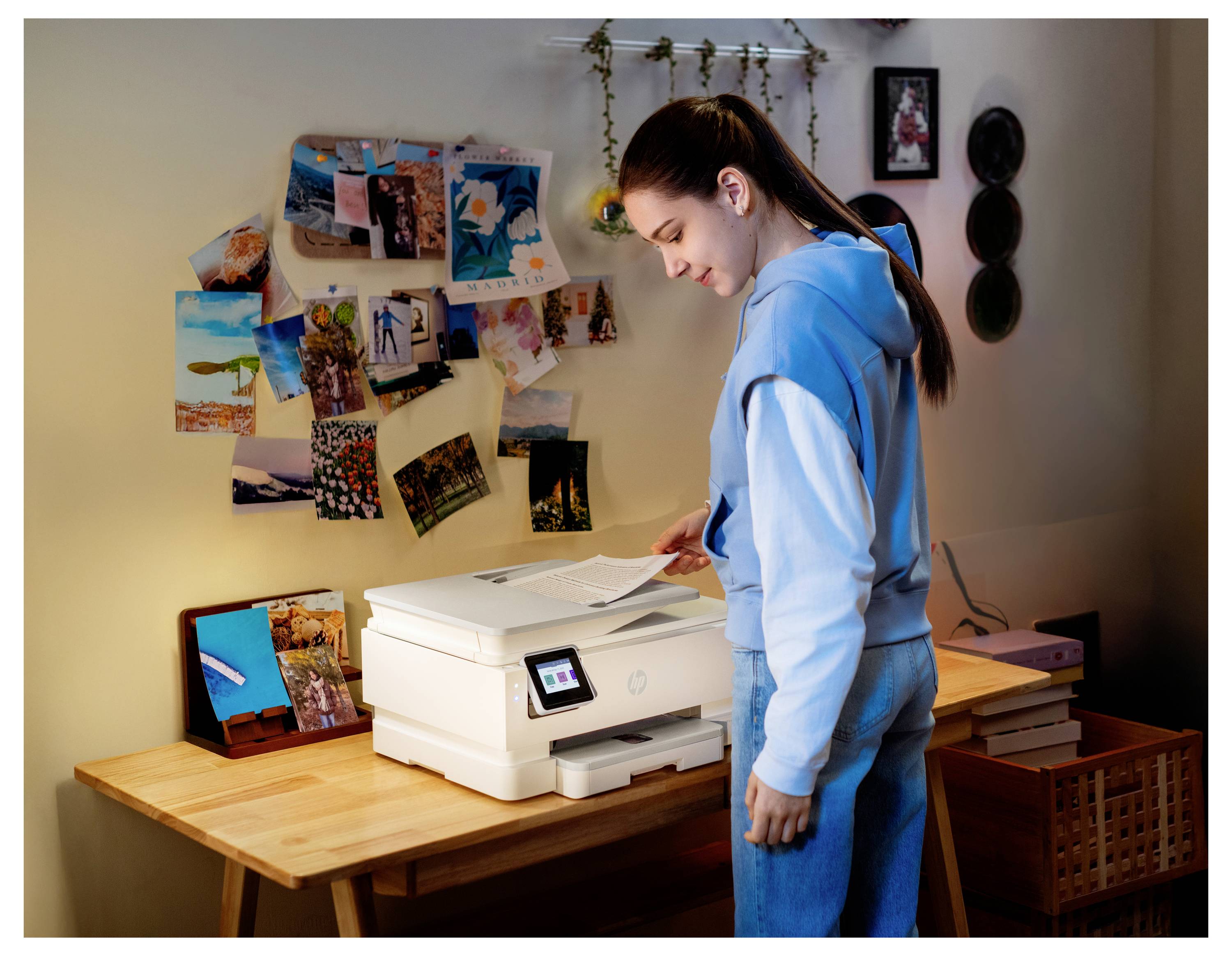 Teenager in blauem Hoodie benutzt einen Drucker an einem Schreibtisch in einem Zimmer mit Fotos und Kunstwerken, die an der Wand angebracht sind.