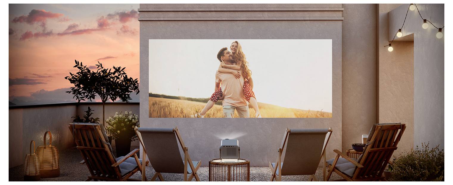 Außenterrasse mit Sonnenuntergang, auf der ein Projektor ein Paar auf einem Feld auf einer großen Leinwand zeigt. Vier Liegestühle sind zur Leinwand ausgerichtet.