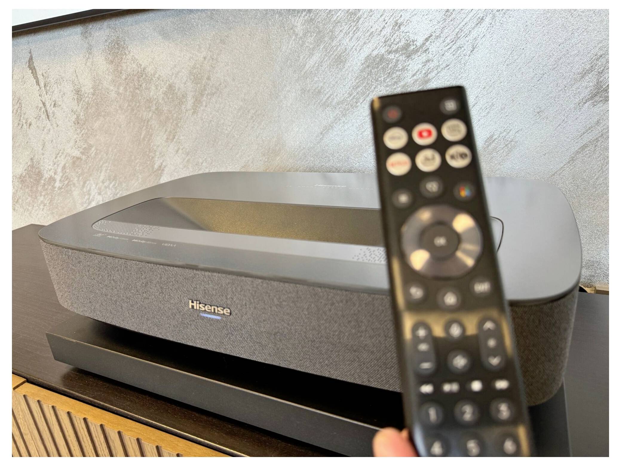 Ein Hisense-Projektor steht auf einem Holztisch, wobei eine Fernbedienung prominent im Vordergrund gehalten wird.