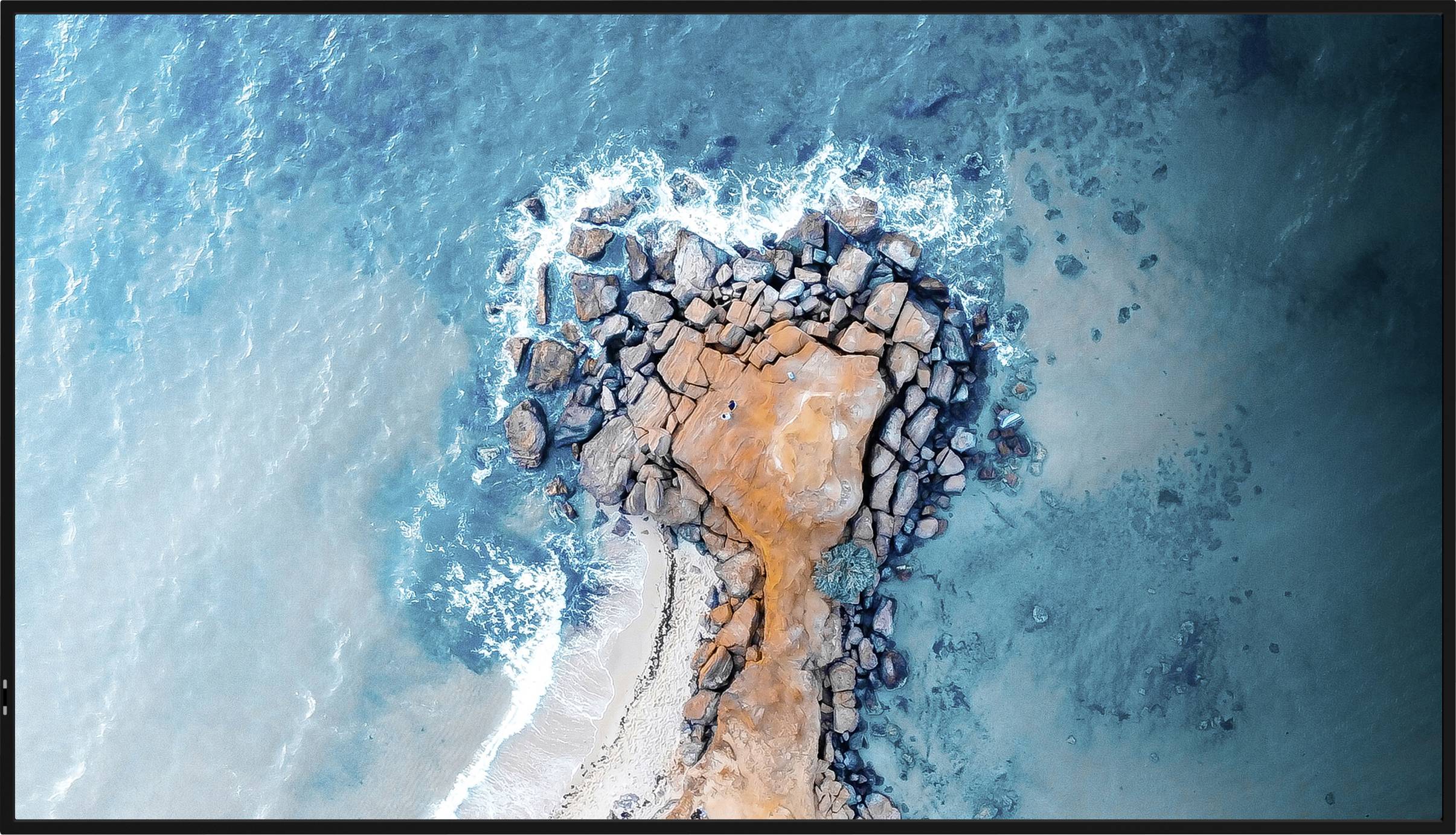 Luftaufnahme einer Felsenformation an der Küste, umgeben von blauen Ozeanwellen, wobei Wasser über die Felsen fließt und einen Kontrast der Texturen schafft.