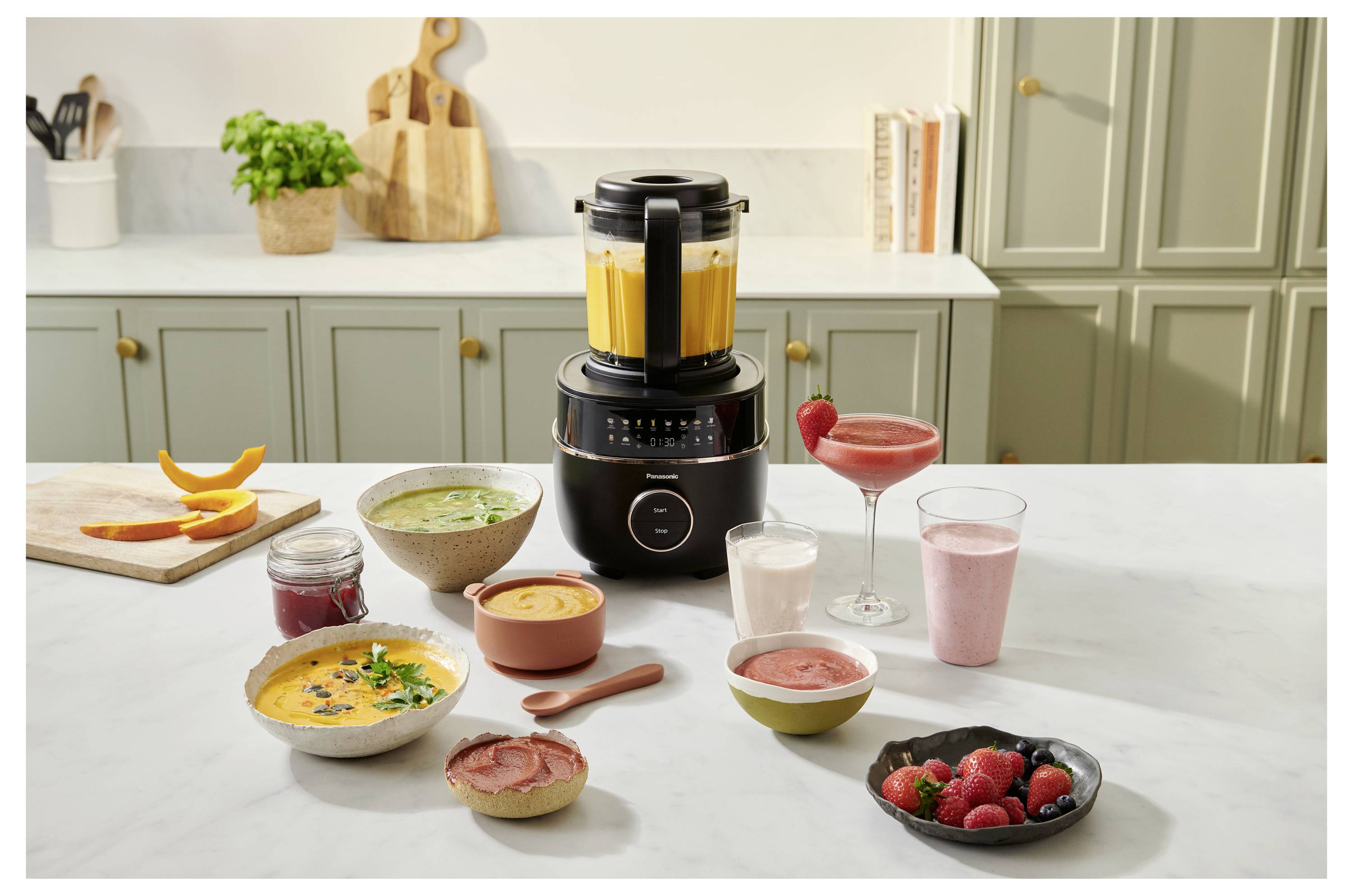 Ein Standmixer auf einer Küchenarbeitsplatte mit verschiedenen Smoothies und Suppen in Schüsseln und Gläsern. Zutaten und ein Schneidebrett befinden sich im Hintergrund.