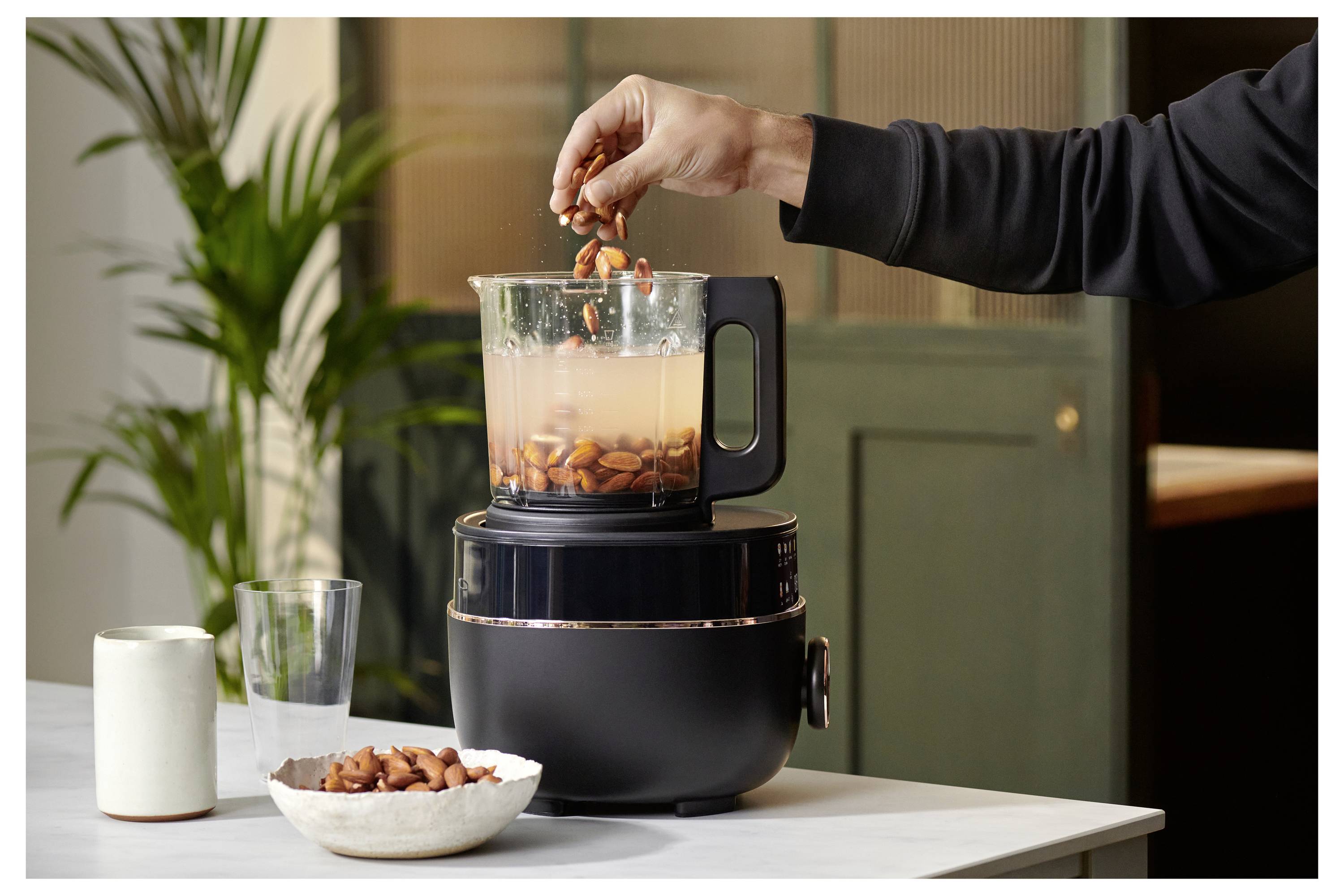 Eine Person gibt Mandeln in einen Mixer, der mit Wasser gefüllt ist und auf einer Küchenarbeitsplatte steht. Eine Pflanze und Küchenutensilien sind im Hintergrund zu sehen.