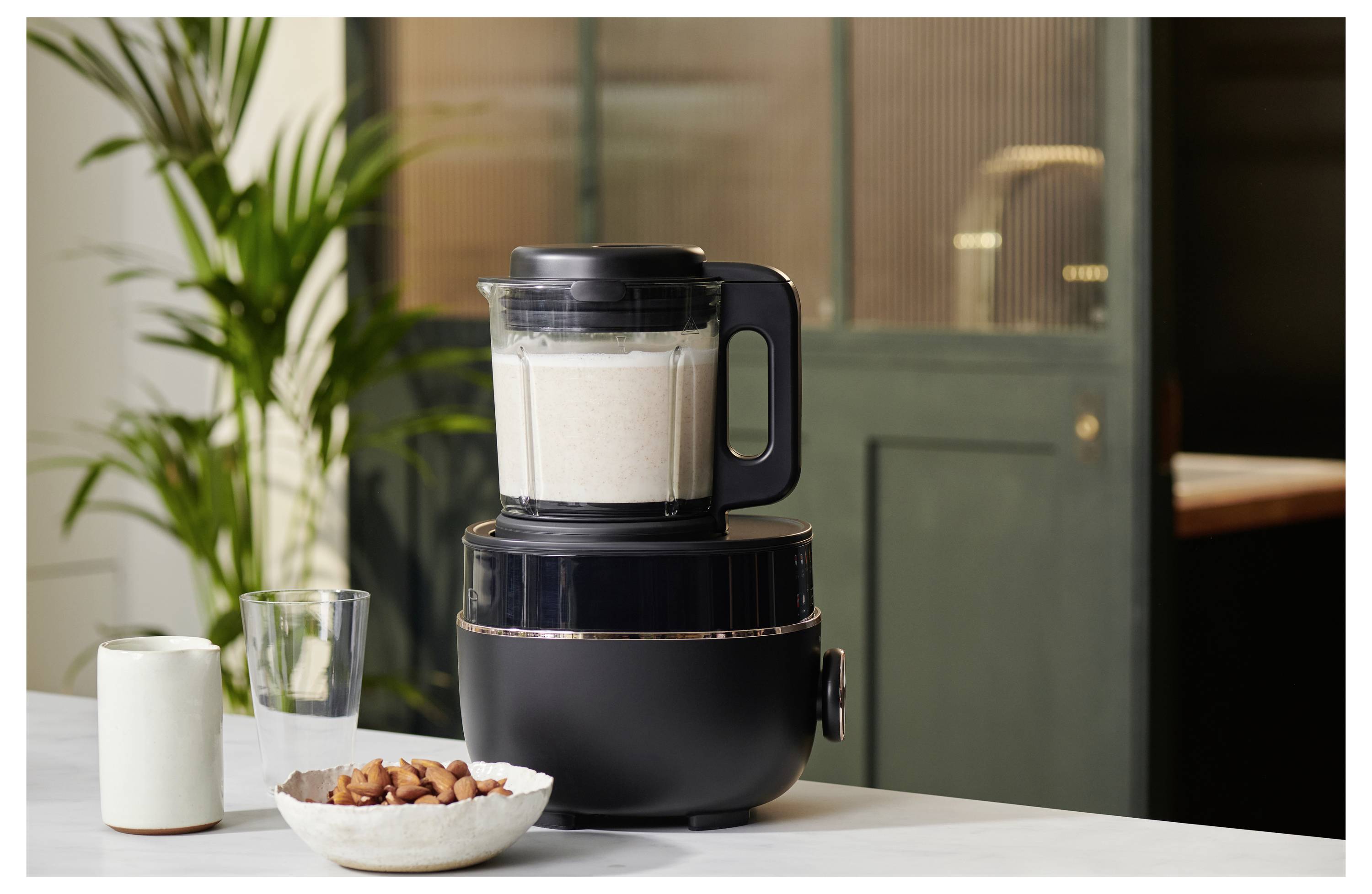 Ein schwarzer Mixer auf einer Küchenarbeitsplatte, gefüllt mit einer weißen Mischung, neben einer Schüssel mit Mandeln und einem Glas, mit einer Pflanze im Hintergrund.