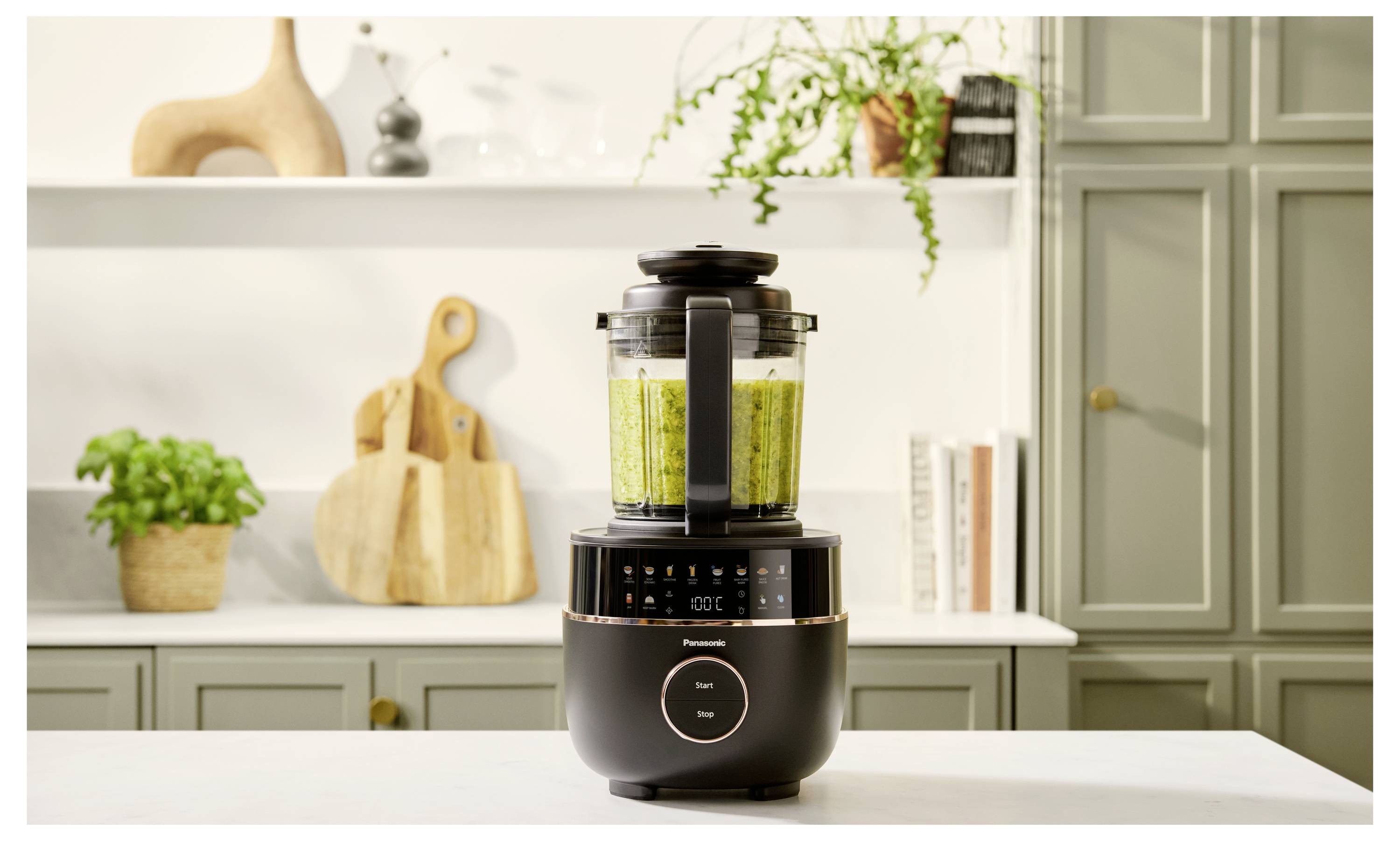Ein Mixer steht auf einer Küchenarbeitsplatte und mixt grüne Zutaten. Im Hintergrund befinden sich Holzutensilien, Pflanzen und Bücher.