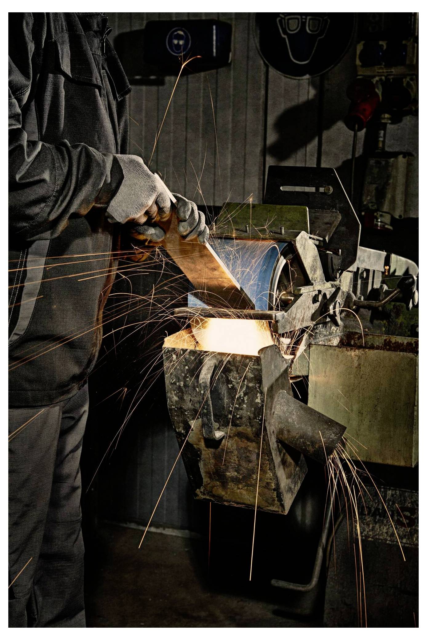 Ein Arbeiter bedient eine Schleifmaschine und erzeugt dabei helle Funken. Er trägt Schutzhandschuhe und eine Jacke und konzentriert sich auf seine Aufgabe in einer industriellen Umgebung.