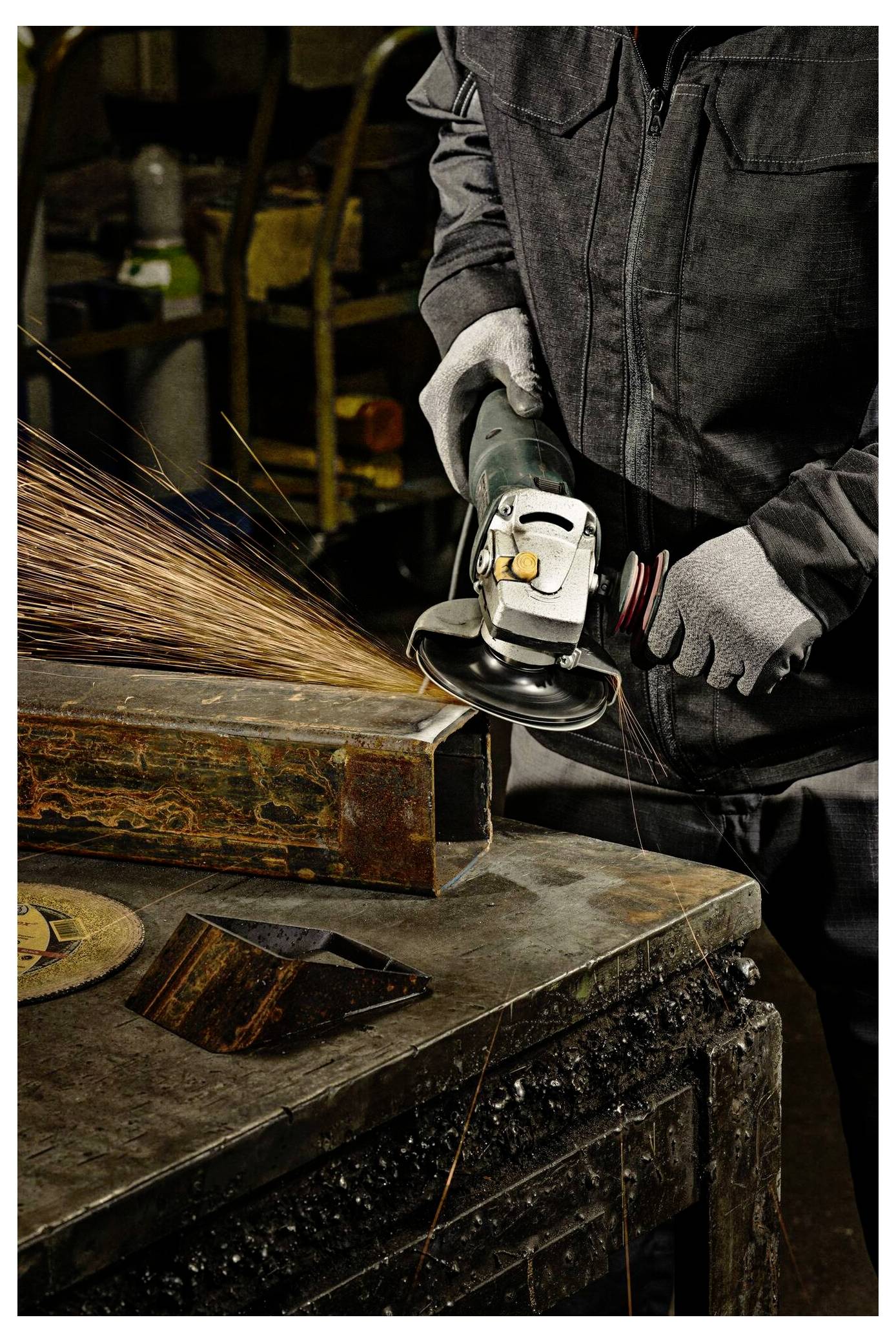 Eine Person mit Handschuhen und einem dunklen Arbeitsanzug benutzt einen Winkelschleifer, der Funken auf einem verrosteten Metallträger in einer Werkstatt emittiert.