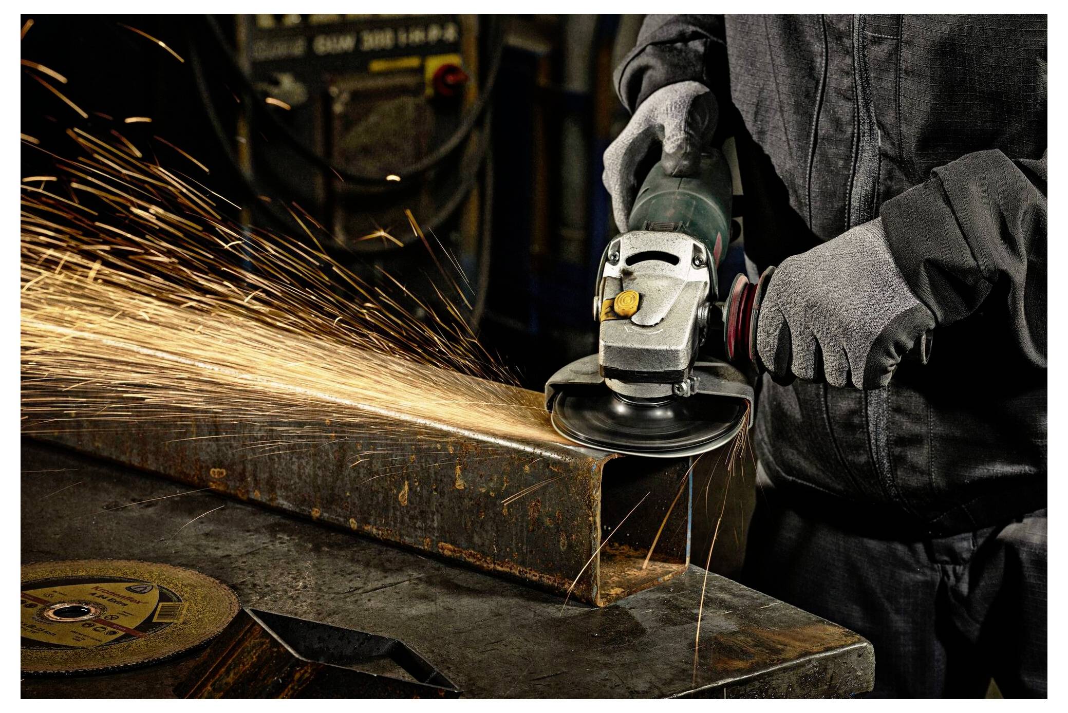 Ein Arbeiter schleift einen Metallträger mit einer Winkelschleifmaschine und erzeugt helle Funken. Der Arbeiter trägt Schutzhandschuhe und eine dunkle Arbeitsuniform.