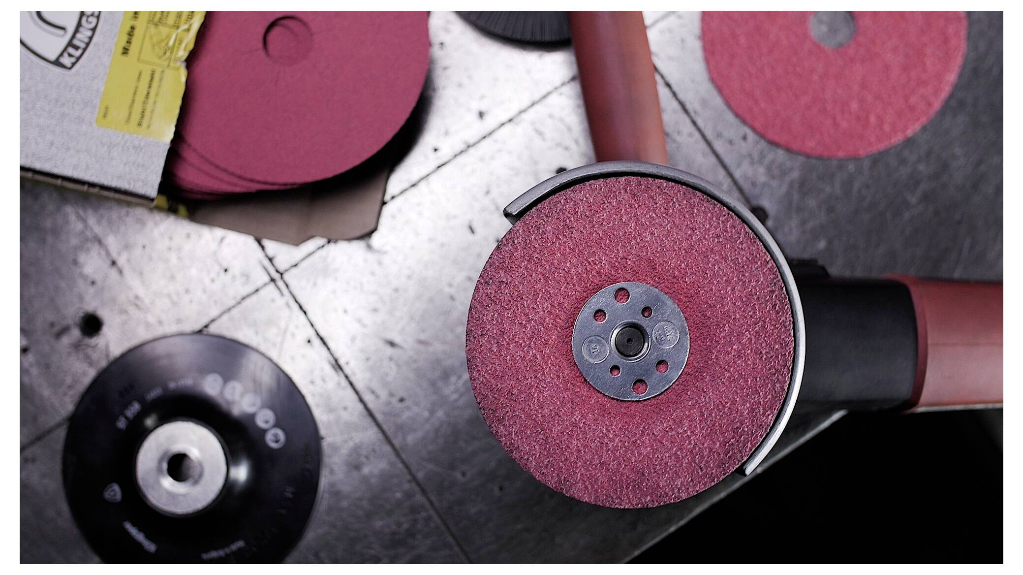 Eine rote Schleifscheibe ist an einer Winkelschleifmaschine befestigt und ist von weiteren Scheiben auf einer metallischen Oberfläche umgeben, was eine Werkstattumgebung andeutet.