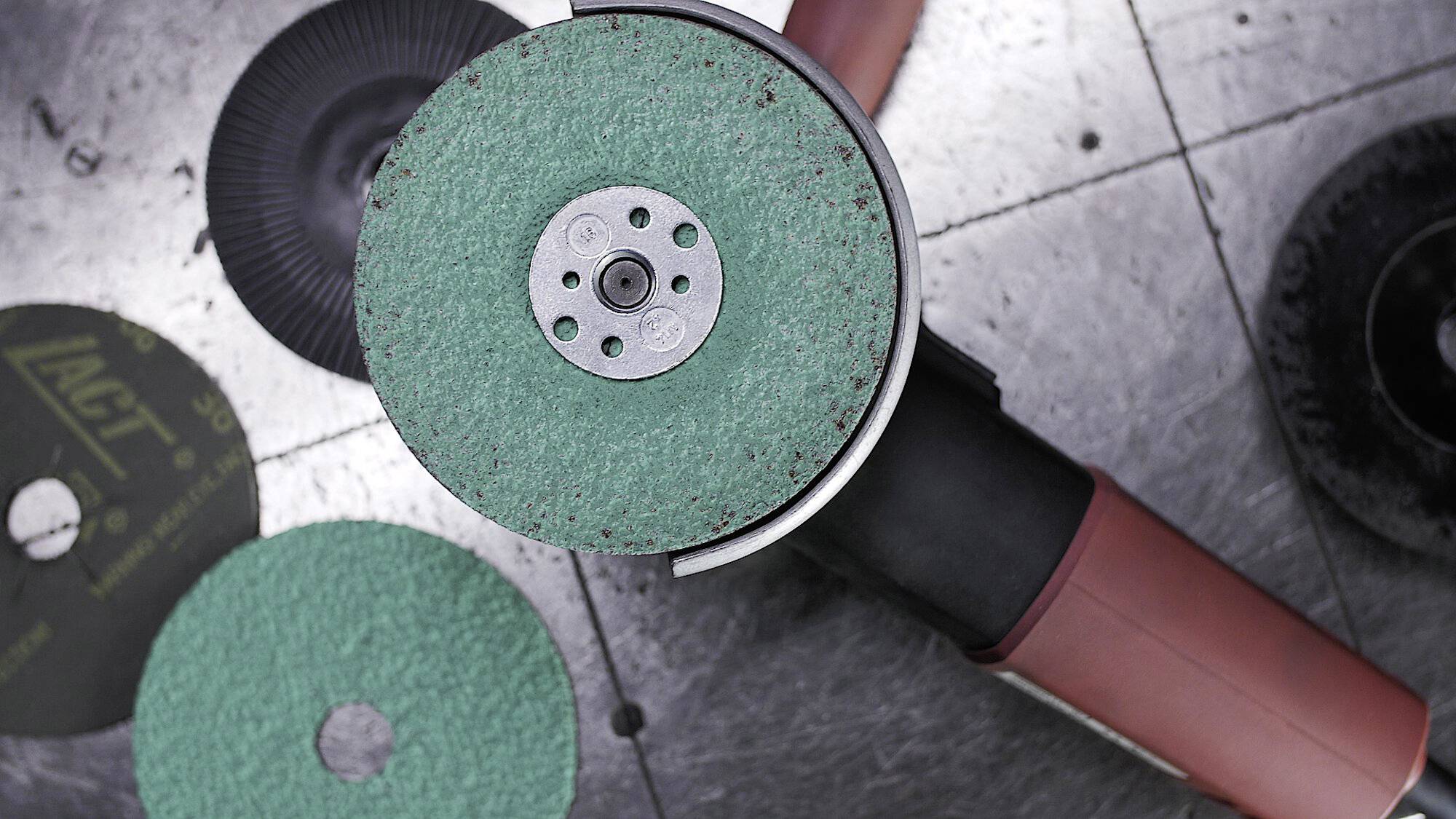 Ein Winkelschleifer mit einer grünen Schleifscheibe, die auf einer Metalloberfläche liegt, umgeben von zusätzlichen Scheiben, was auf industrielle Nutzung hindeutet.