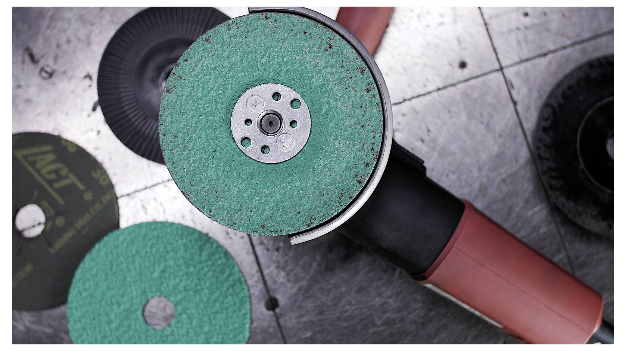 Eine gewinkelte elektrische Schleifmaschine mit einer grünen Schleifscheibe steht auf einer gefliesten Metalloberfläche und wird von zusätzlichen Scheiben begleitet.