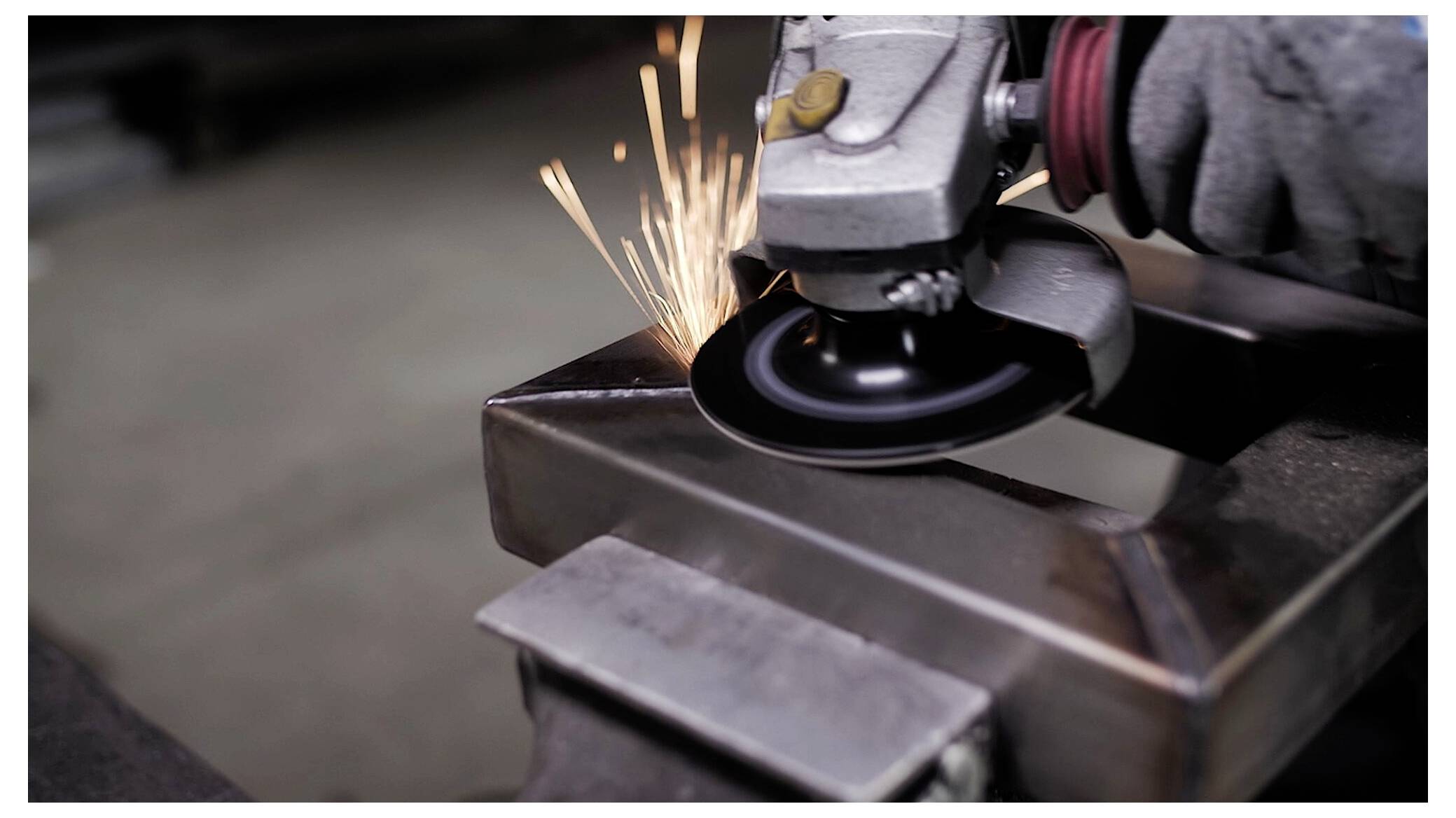 Eine Nahaufnahme einer Winkelschleifer, gehalten von einer Person mit Handschuhen, die beim Schneiden von Metall auf einer Werkbank Funken auswirft.