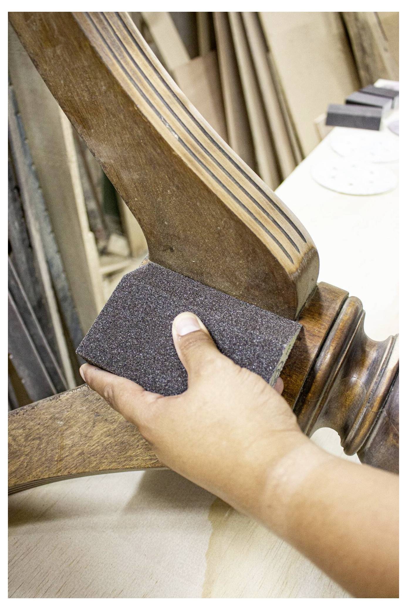 Eine Hand einer Person schleift ein Stuhlbein mit einem Schleifklotz in einer Werkstatt, umgeben von Holzmaterialien und Werkzeugen.