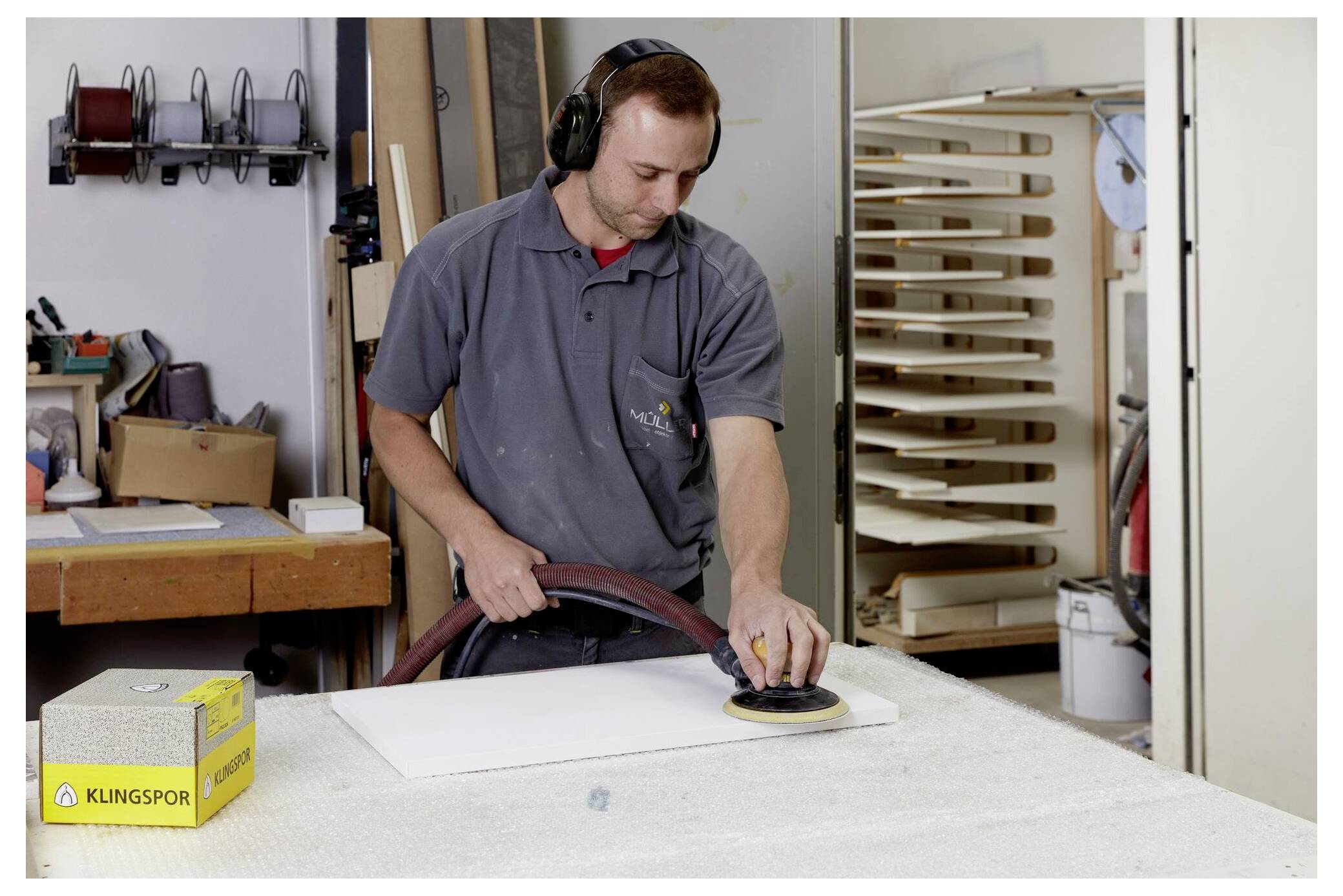 Person in einer Werkstatt, die mit einer Maschine eine weiße Tafel schleift; trägt Gehörschutz und einen grauen Overall. Werkbank und Werkzeuge im Hintergrund.