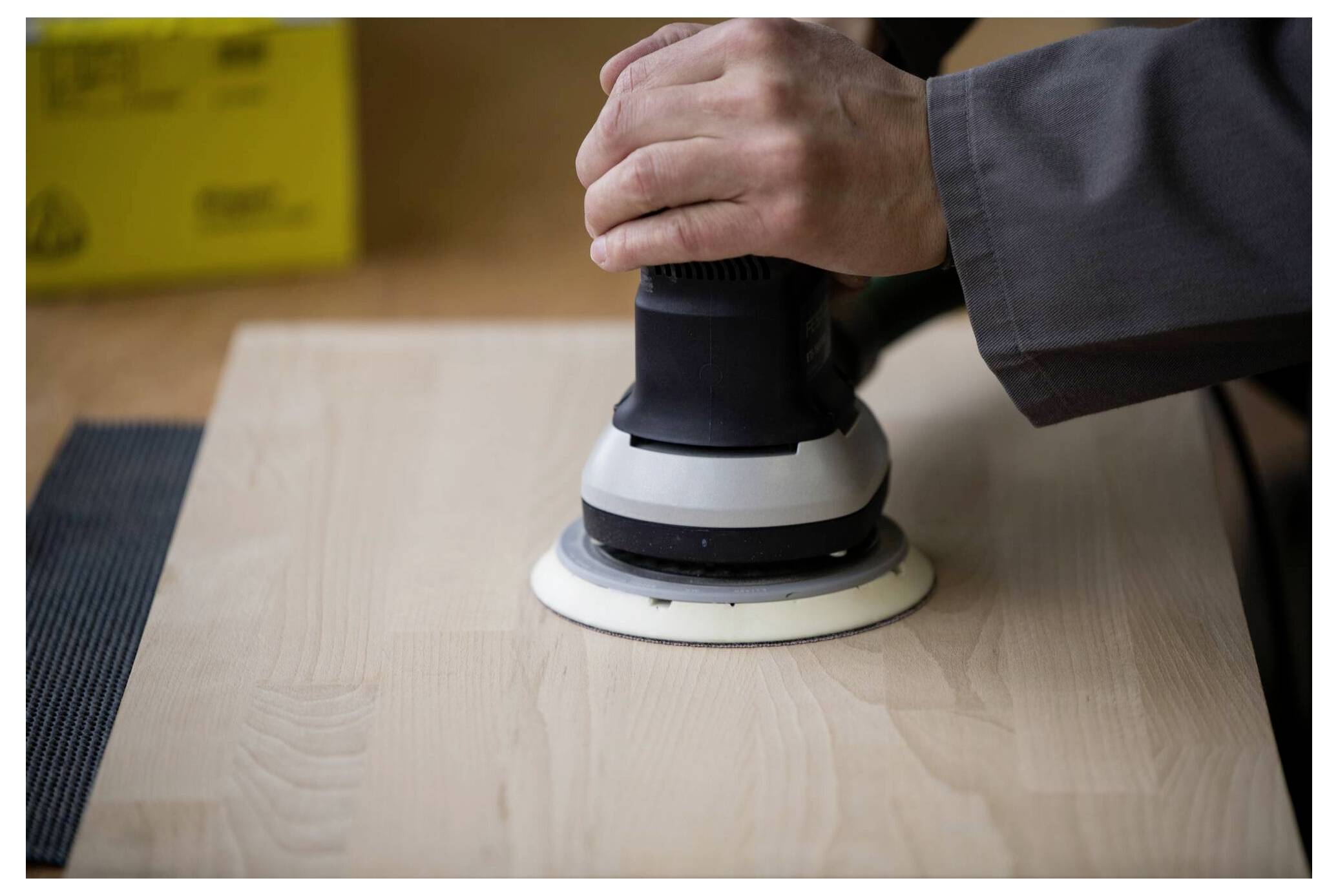 Eine Person verwendet einen Elektrohobel auf einer Holzoberfläche und glättet sie. Sie trägt einen grauen Ärmel, was auf eine professionelle oder industrielle Umgebung hindeutet.