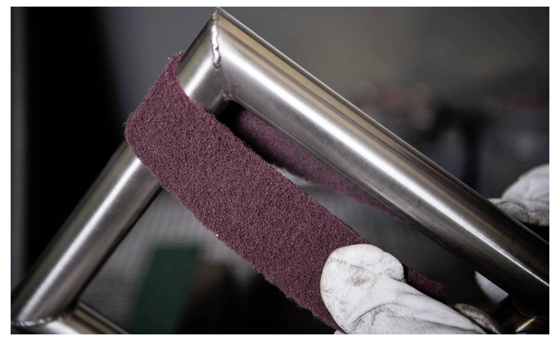 Eine Person verwendet ein marronefarbenes Schleifpad, um einen Edelstahlrahmen zu polieren und hebt dabei die glatte, glänzende Oberfläche des Metalls hervor.