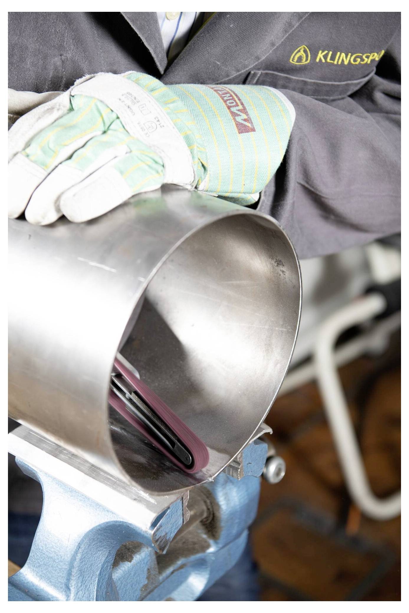 Eine Person mit Handschuhen arbeitet an einem Metallrohr mit einem Werkzeug. Die Werkstattumgebung deutet auf eine Konzentration auf Metallbearbeitung oder Reparaturen hin.