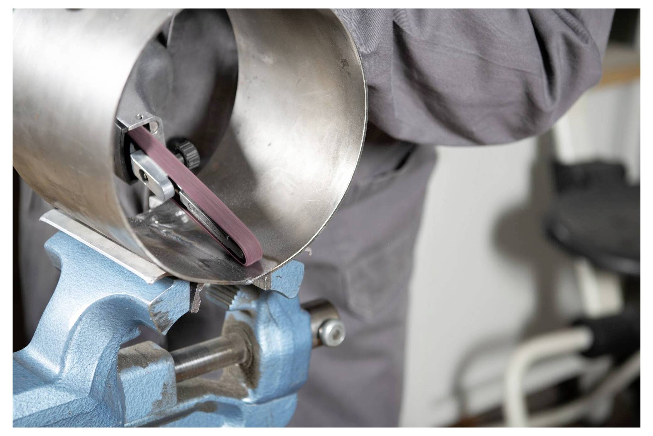 Person, die einen Metallschleifer an einem zylindrischen Objekt verwendet, das in einem blauen Schraubstock eingespannt ist und einen grauen Arbeitsanzug trägt. Fokus auf den Schleifprozess.