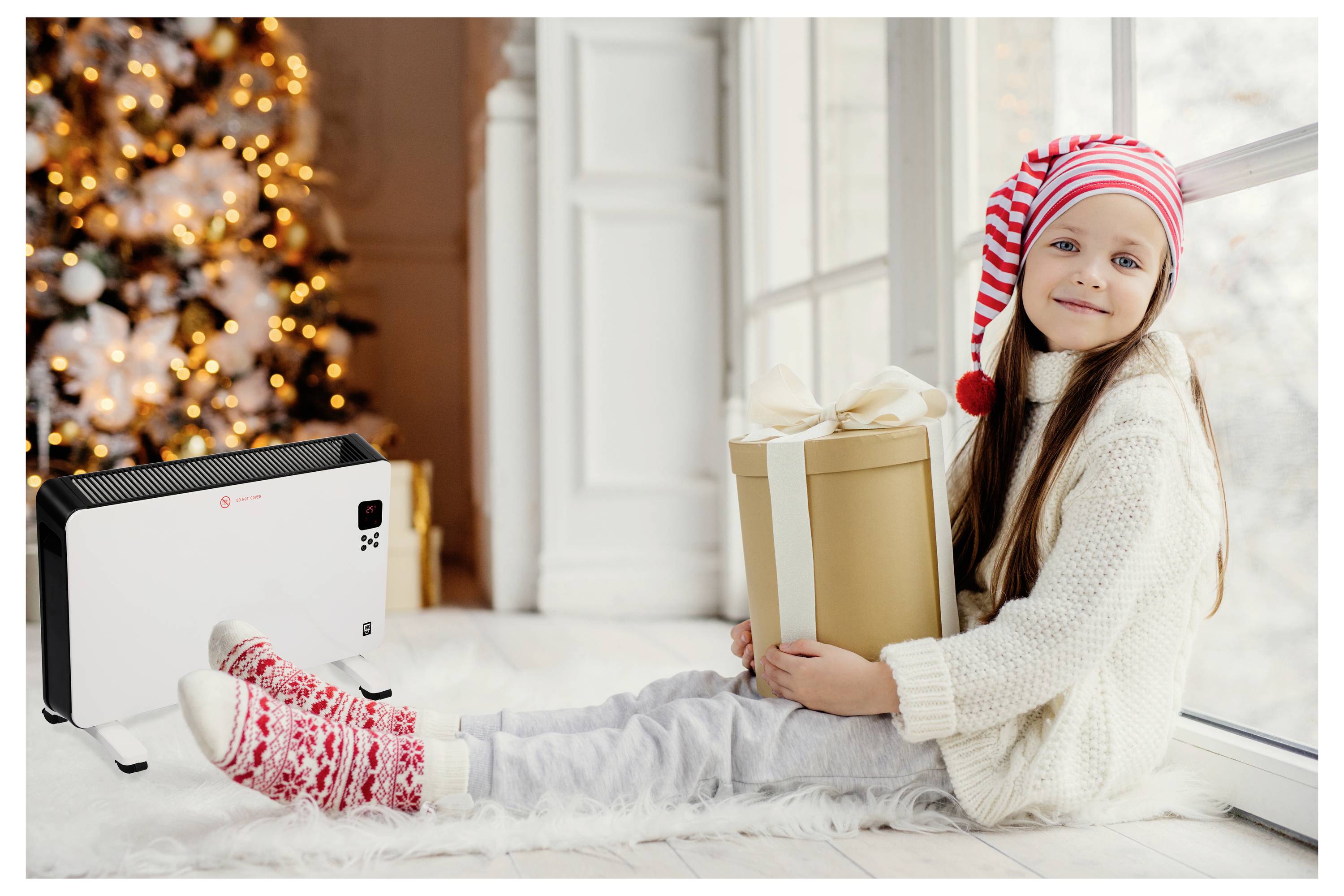 Ein Kind mit einer festlichen Mütze und einem Pullover sitzt an einem Fenster und hält ein verpacktes Geschenk. Ein dekorativer Heizkörper und ein Weihnachtsbaum mit Lichtern befinden sich im Hintergrund.