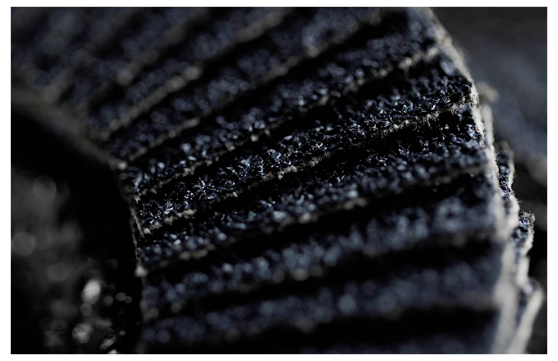 Nahaufnahme einer strukturierten, dunkelgrauen Oberfläche mit parallelen Rillen, möglicherweise ein Stoff oder ein Industriematerial, mit Betonung auf Textur und Details.