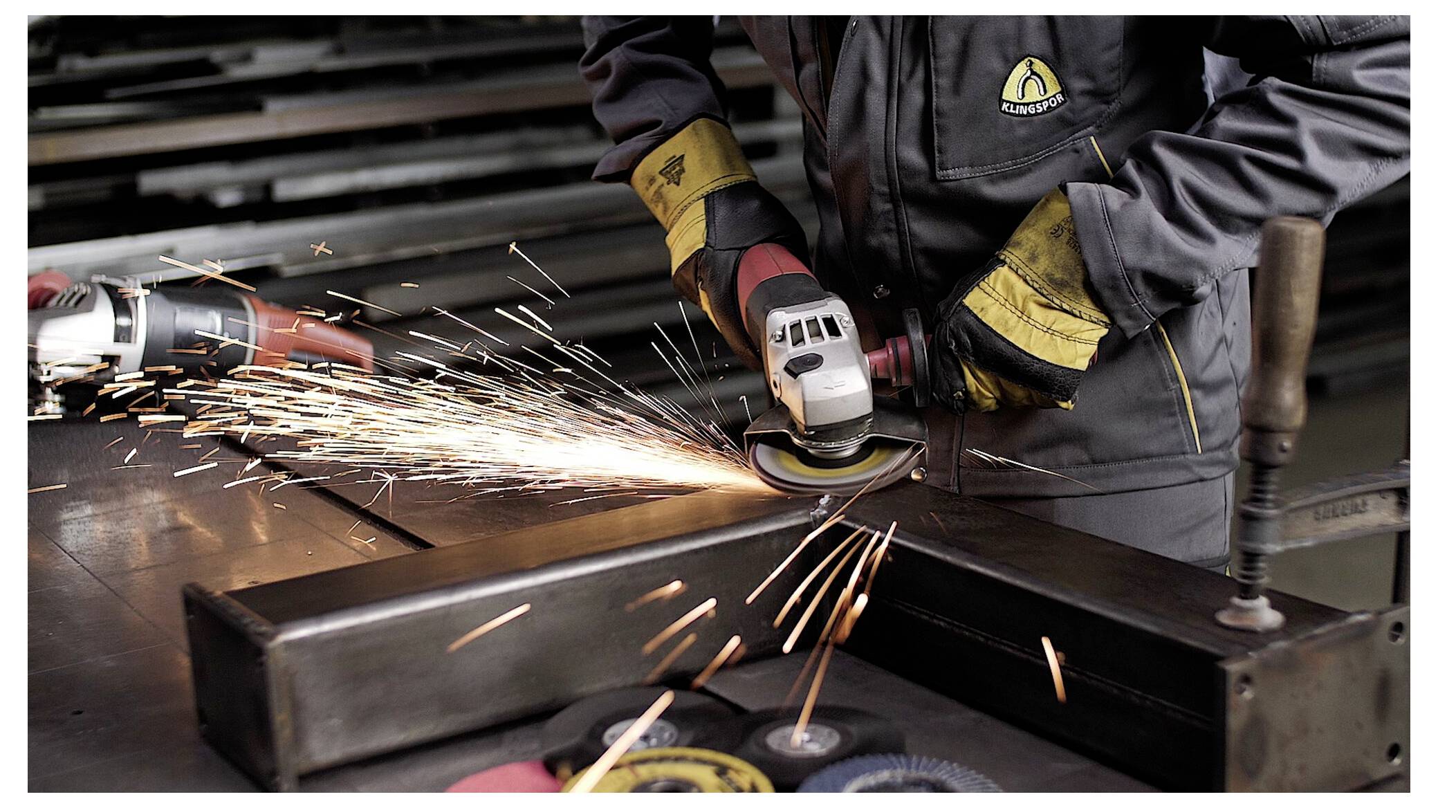 Eine Person in Schutzausrüstung benutzt einen Winkelschleifer an einem Metallstück, wobei Funken in einer Werkstattumgebung entstehen.