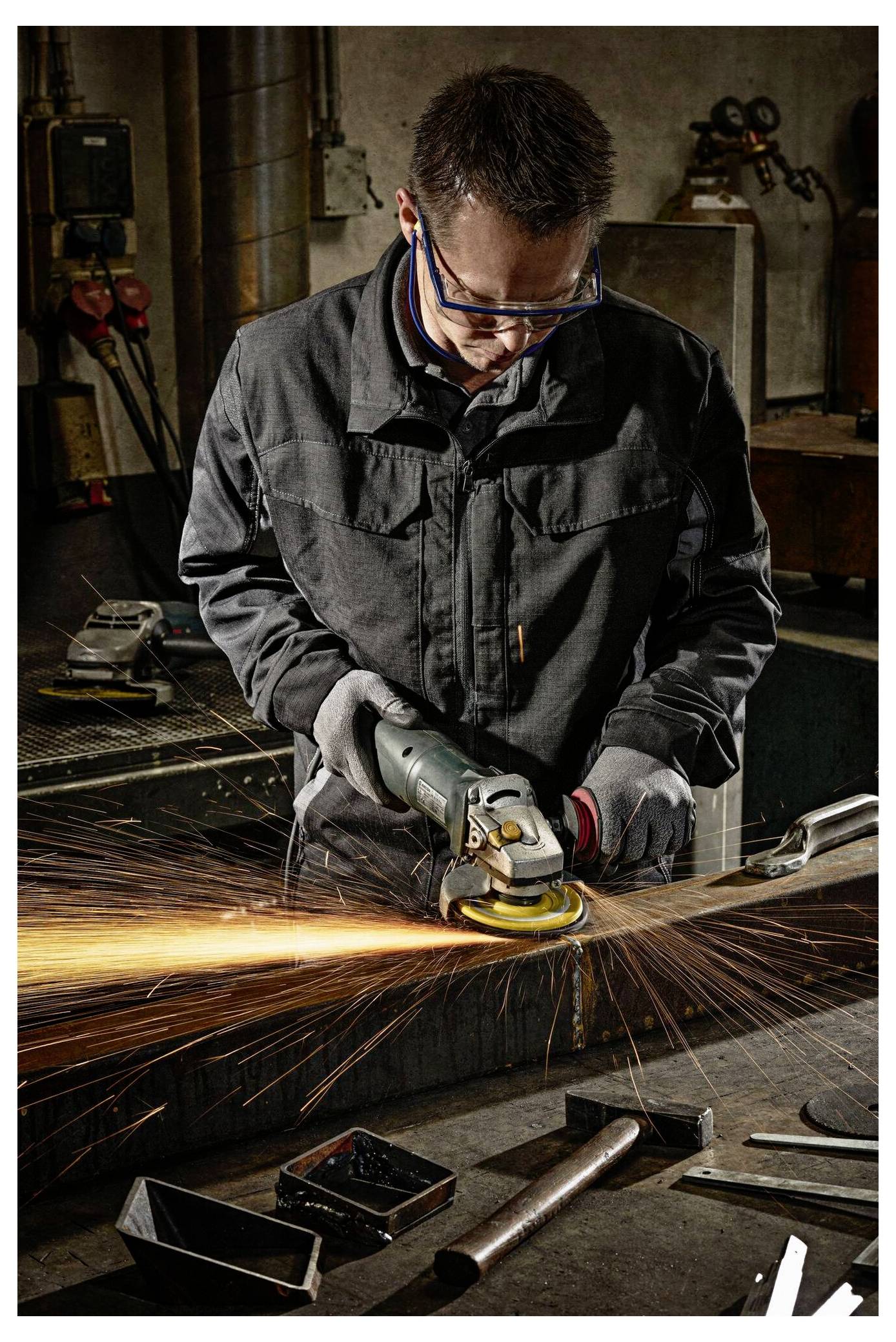 Eine Person mit Sicherheitsausrüstung verwendet einen Winkelschleifer auf einer Metalloberfläche in einer Werkstatt und erzeugt dabei helle orangefarbene Funken. Im Hintergrund befinden sich Werkzeuge und Ausrüstungsgegenstände.