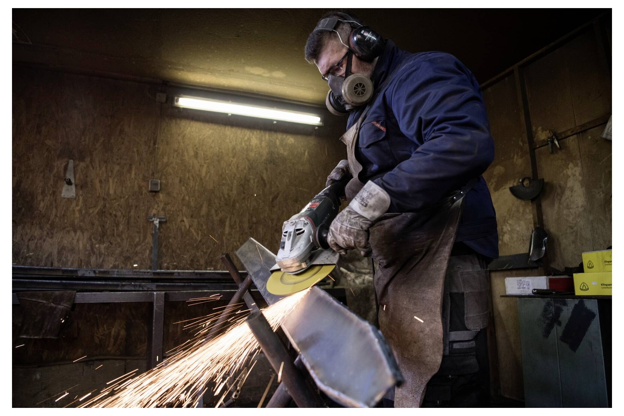 Eine Person in Schutzausrüstung verwendet einen Winkelschleifer an einem Metallobjekt und erzeugt Funken in einer Werkstattumgebung.