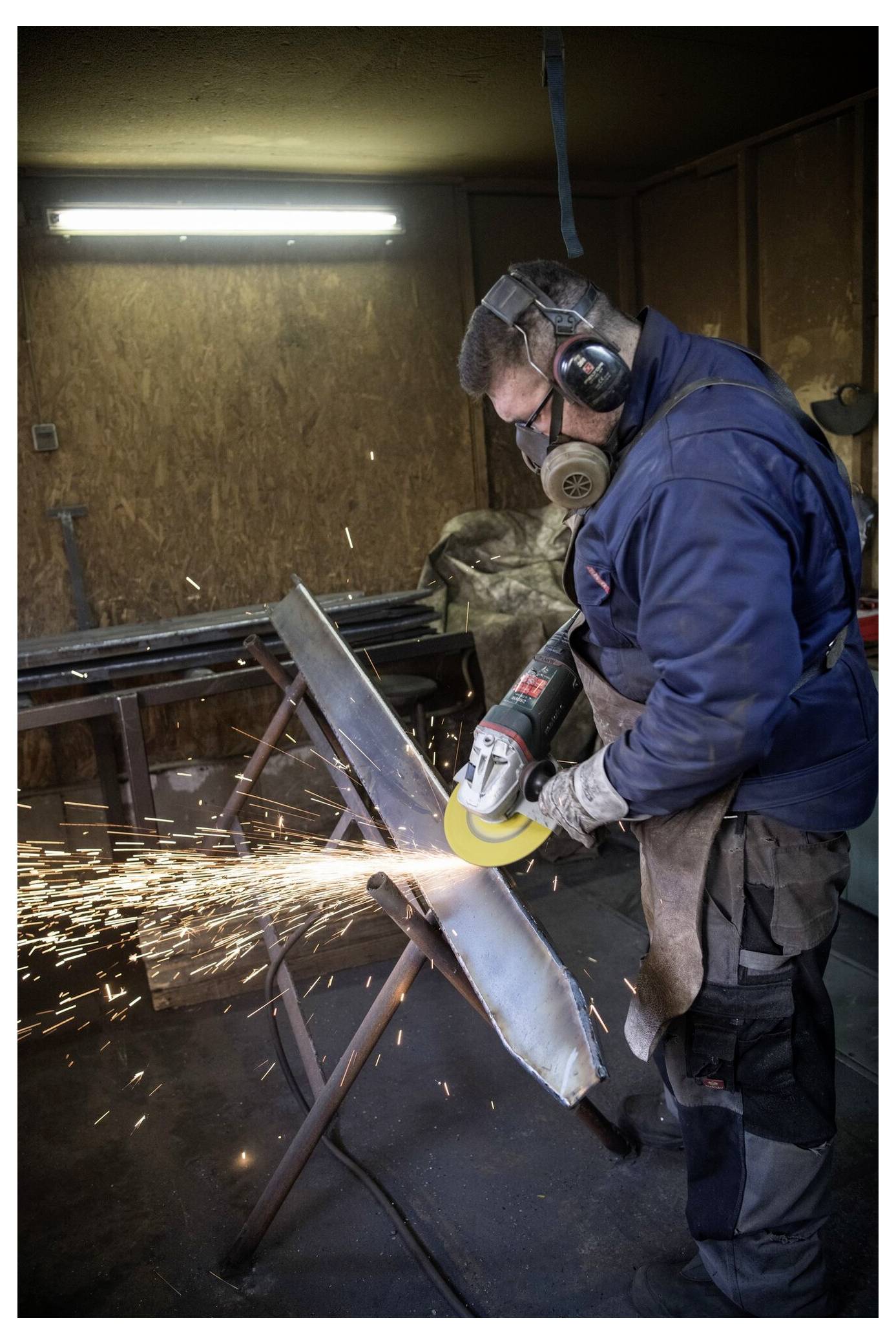 Eine Person in Schutzausrüstung benutzt ein Schleifwerkzeug an einem Metallstück, wobei Funken in einer Werkstattumgebung sprühen.