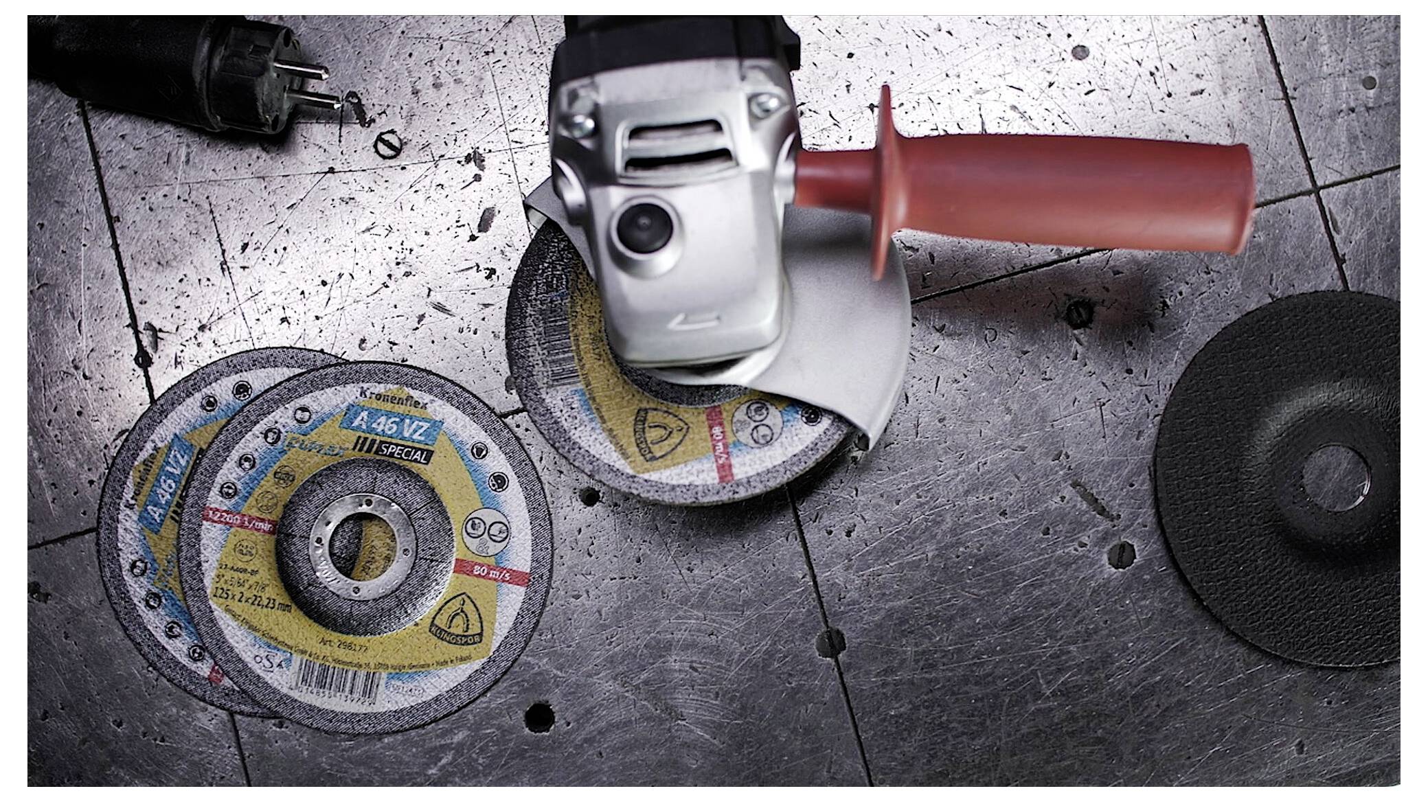 Eine Winkelschleifmaschine liegt auf einer Metalloberfläche mit zwei Trennscheiben in der Nähe, eine angebracht und eine abgenommen. Ein Stromstecker ist oben links sichtbar.