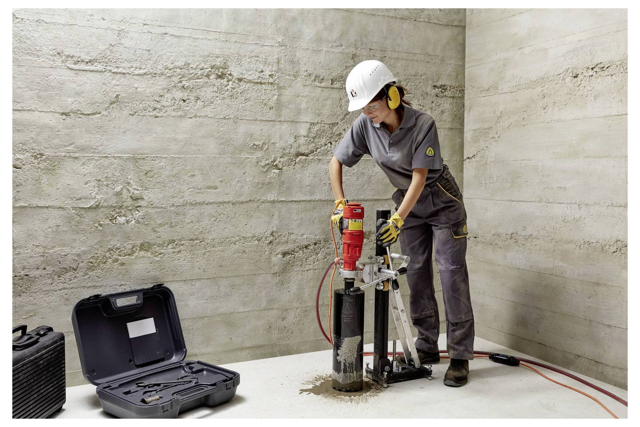 Eine Person in Arbeitskleidung bedient eine Kernbohrmaschine auf einem Betonboden. Werkzeuge und ein geöffneter Koffer befinden sich in der Nähe. Die Umgebung wirkt industriell.