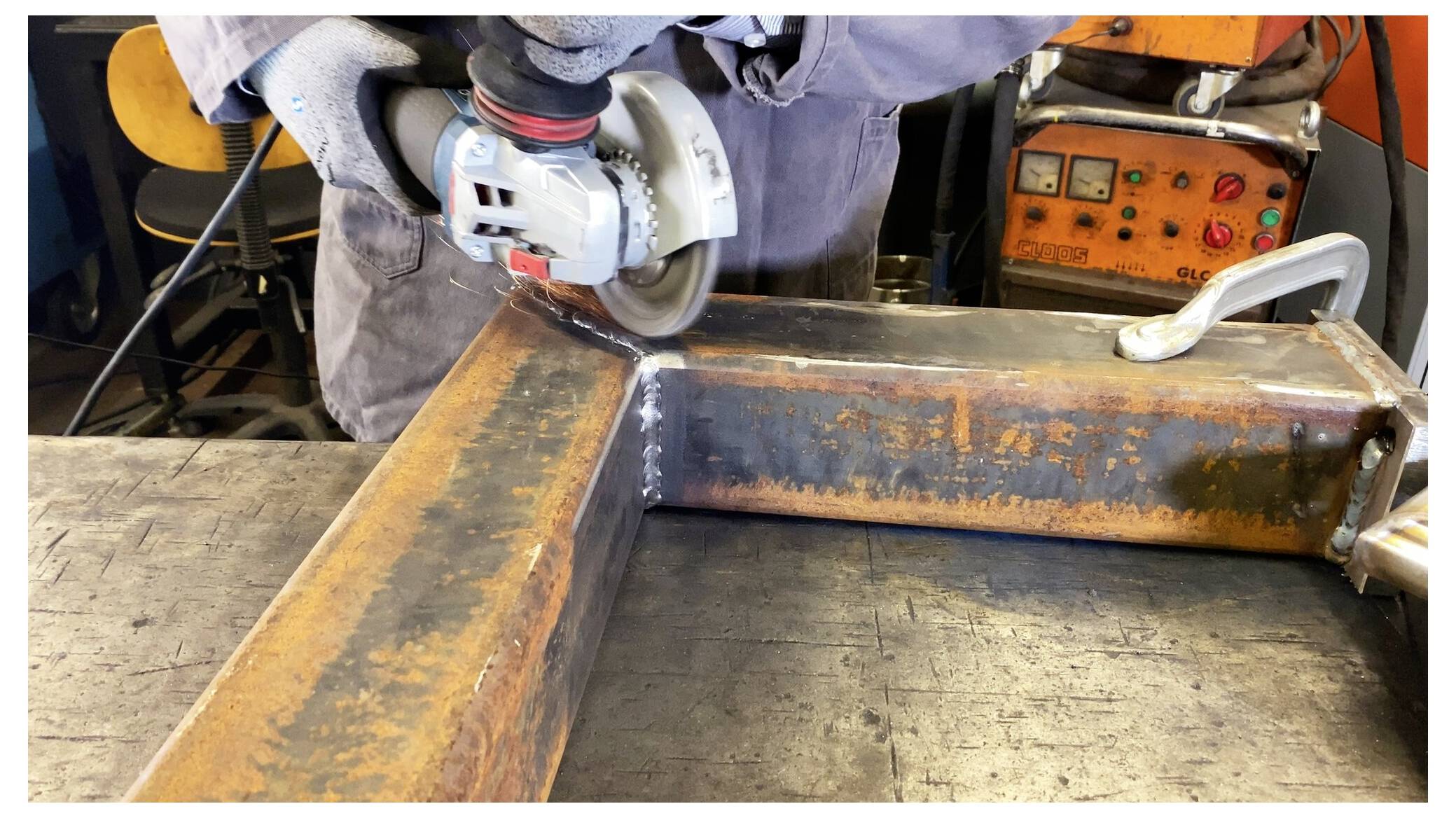 Eine Person mit Handschuhen verwendet einen Winkelschleifer, um die Kanten von zwei verbundenen Metallträgern zu glätten. Im Hintergrund ist ein Bedienfeld zu sehen.