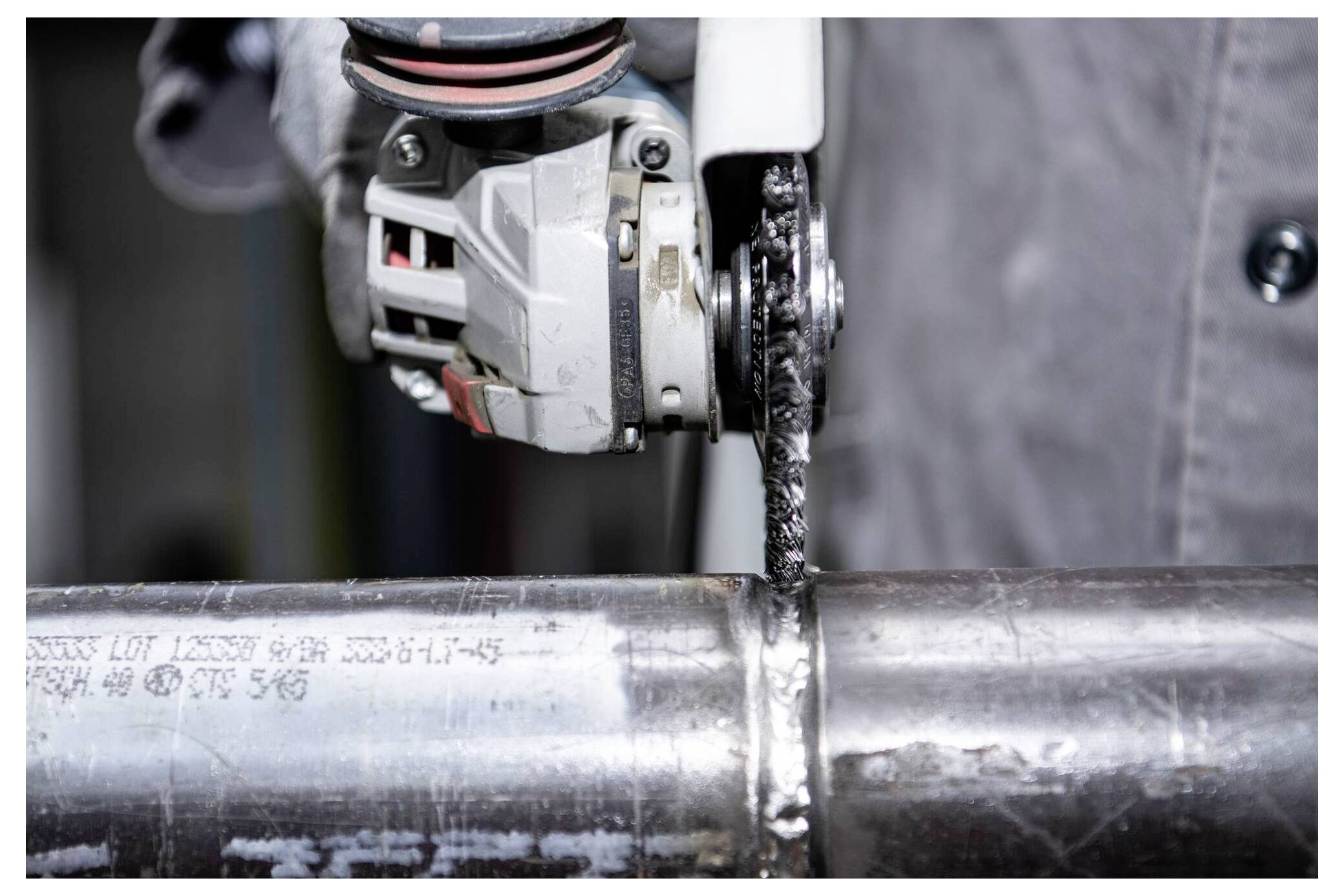 Ein Arbeiter verwendet eine Handwinkelschleifmaschine, um ein Metallrohr zu schneiden. Das Werkzeug sendet Funken aus, die auf aktives Schneiden hinweisen. Das Rohr trägt gravierte Schrift.