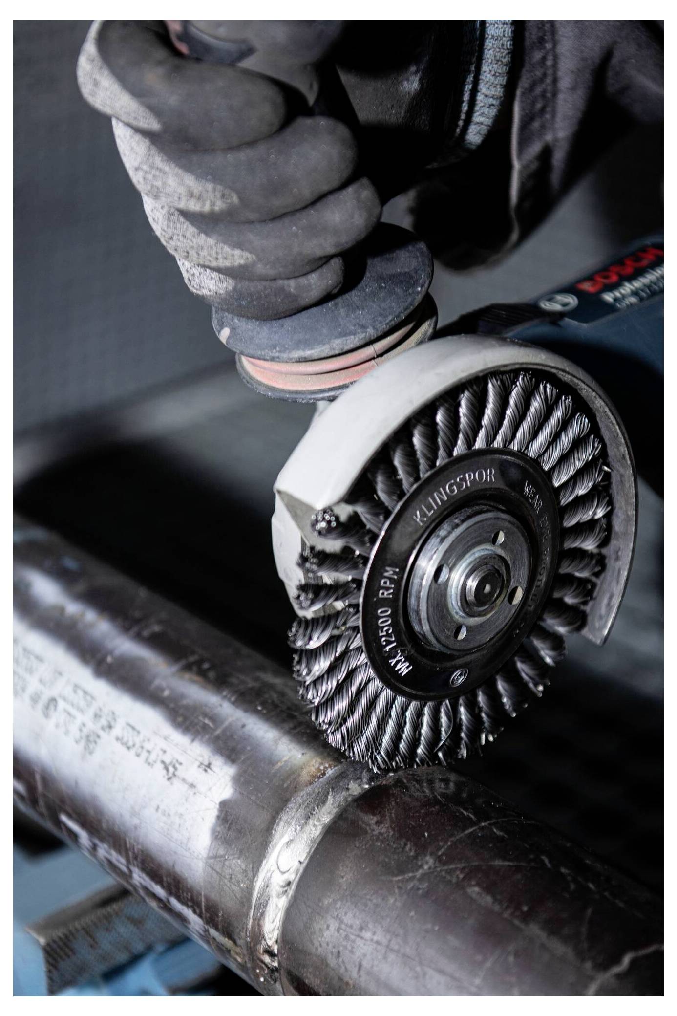 Eine Hand in einem Handschuh verwendet einen Winkelschleifer mit einer Drahtbürsten-Aufsatz, um die Oberfläche eines Metallrohrs zu reinigen oder zu polieren und damit industrielle Arbeit zu veranschaulichen.
