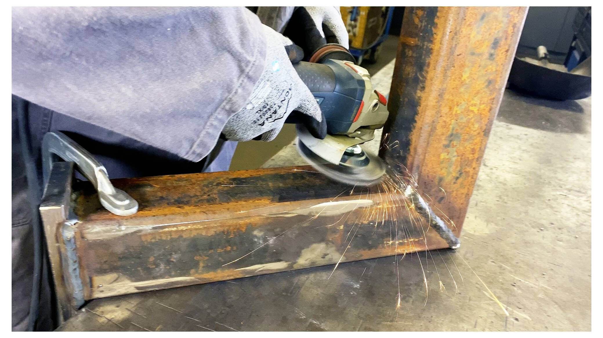 Eine Person mit Handschuhen verwendet einen Winkelschleifer an einem Metallrahmen und erzeugt dabei Funken. Die Arbeit scheint in einer Werkstatt stattzufinden.