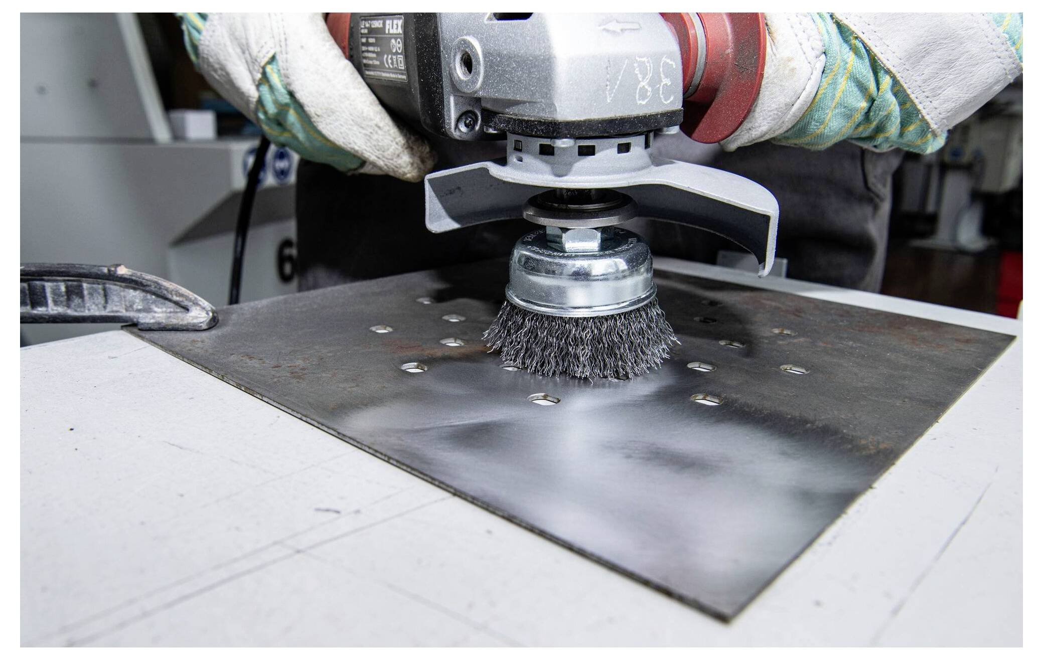 Eine Person verwendet eine Schleifmaschine mit einer Drahtbürsten-Aufsatz, um ein Metallblech zu reinigen. Sie trägt Schutzhandschuhe und arbeitet in einem industriellen Umfeld.