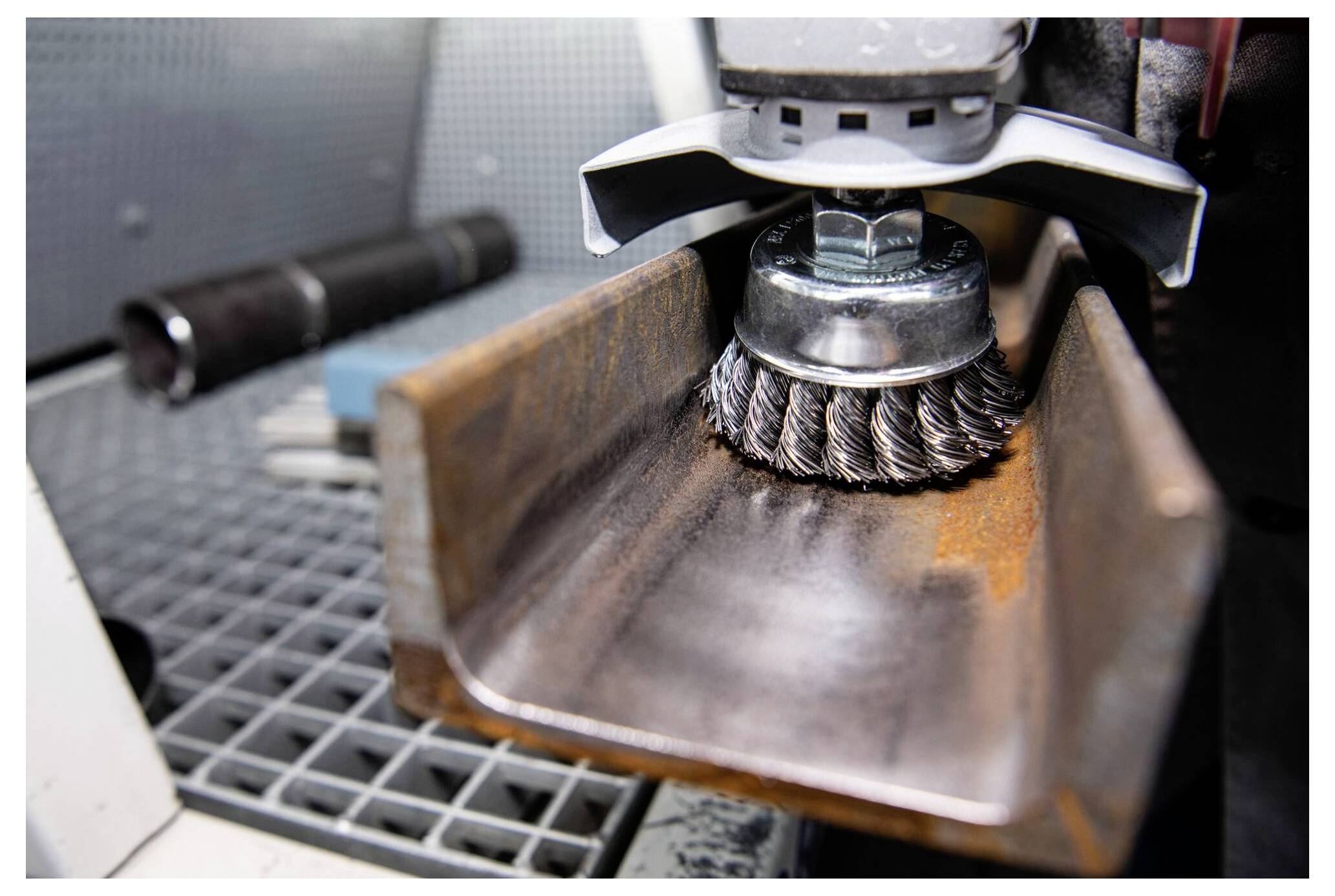 „Eine Nahaufnahme eines Elektrowerkzeugs mit einer Drahtbürsten-Aufsatz, der Rost von einem Metallträger in einer Werkstattumgebung reinigt.