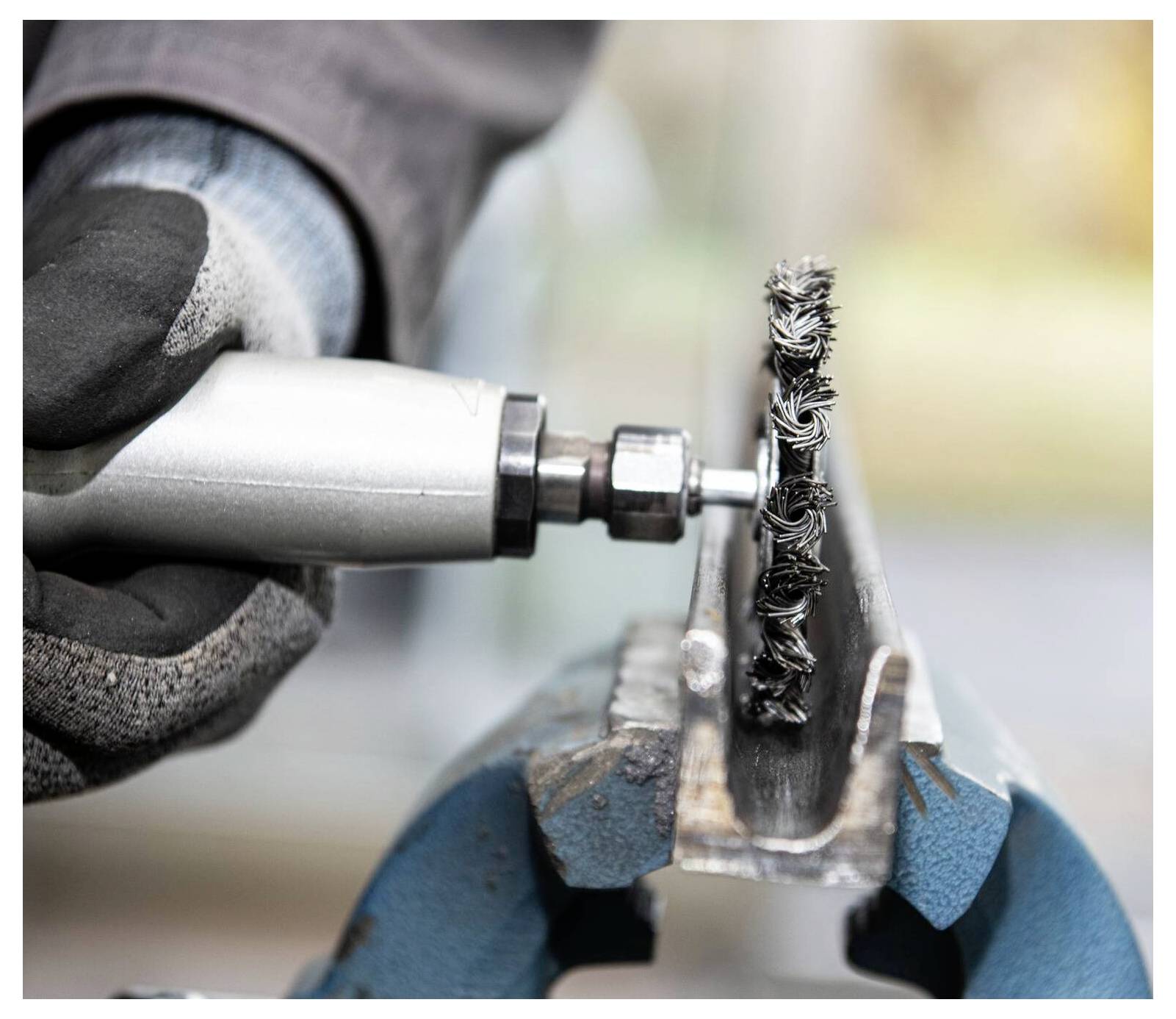 Eine Handschuh tragende Hand verwendet ein Rotationswerkzeug mit Drahtbürsten-Aufsatz, um eine in einem blauen Schraubstock befestigte Metallkante zu reinigen und deutet damit auf eine Werkstattumgebung hin.